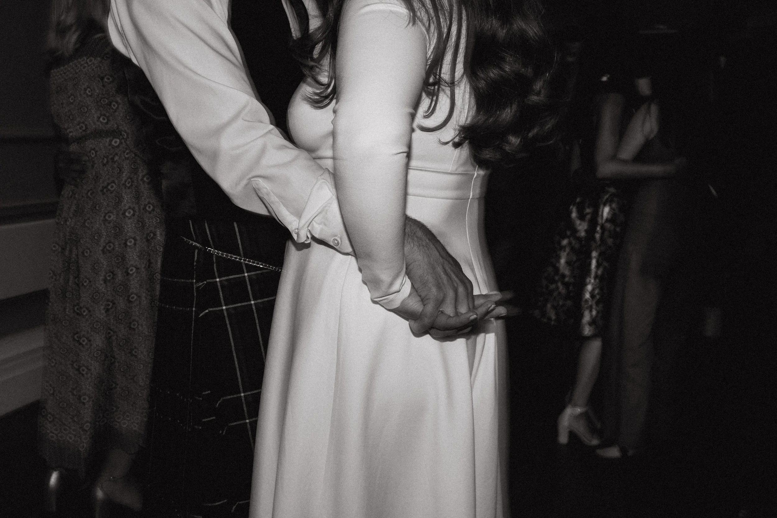 A black and white photo of a woman in a white dress with long dark hair, holding hands with a man in a patterned skirt and a light-colored shirt, in a dimly lit setting with other people in the background.