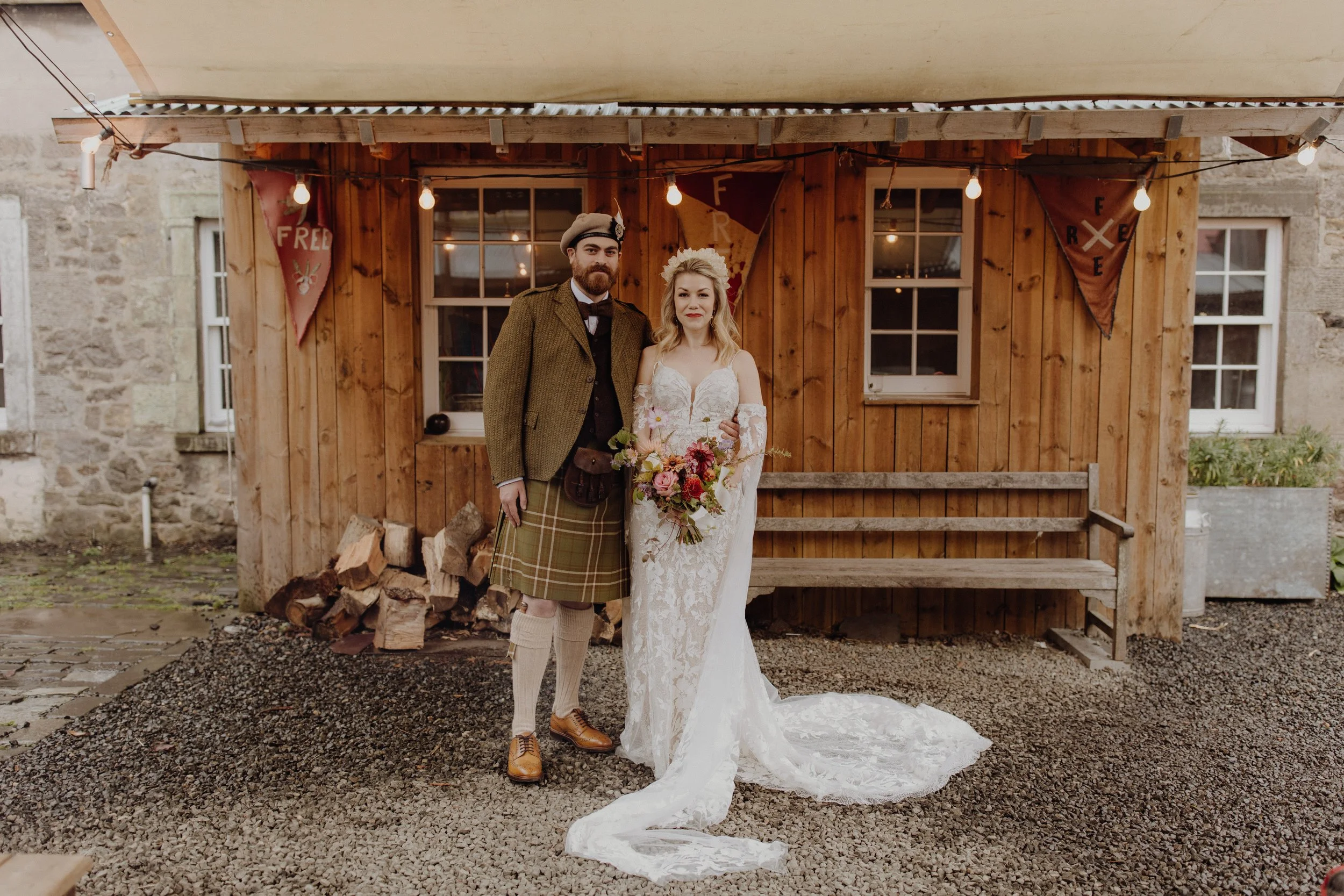 Bride and groom standing under a tent in front of a wooden shed at the free company wedding venue in Edinburgh, With bride in whimsical dress with floor lenght sleeves and groom in traditional kilt