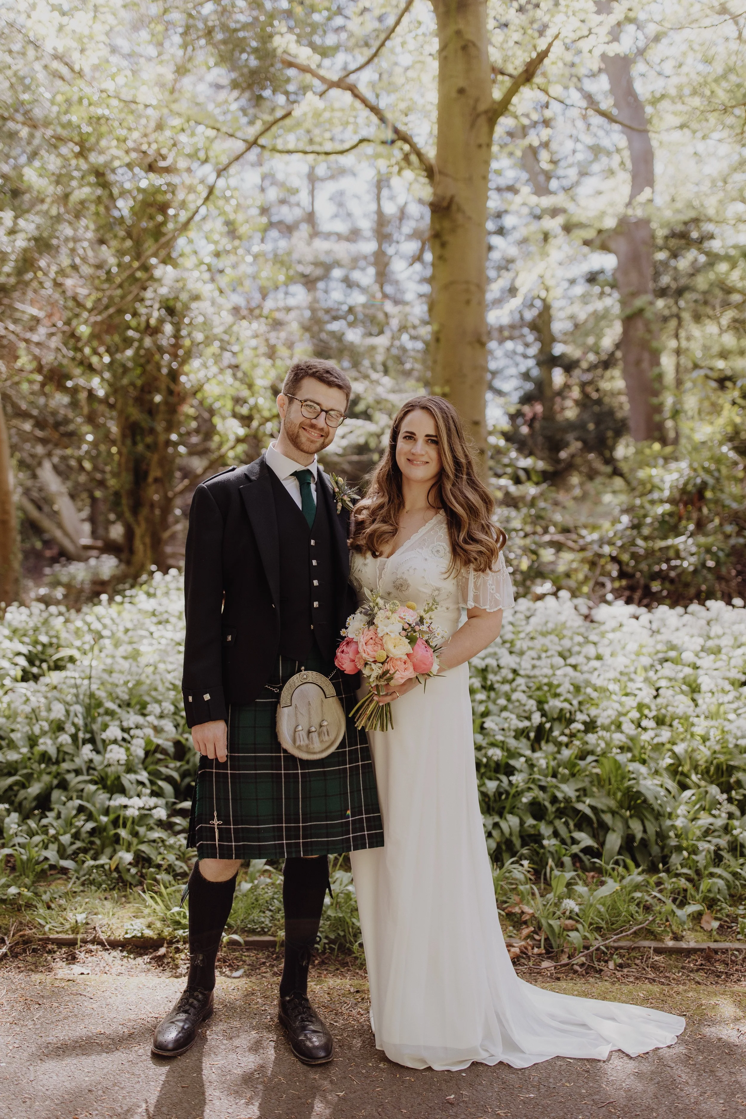 Romantic outdoor portrait at Netherbyres House of wedding couple standing in a sunlit woodland filled with blooming wild garlic with groom wearing traditional kilt and sporran and bride in modest white dress with satin and lace pink white bouquet