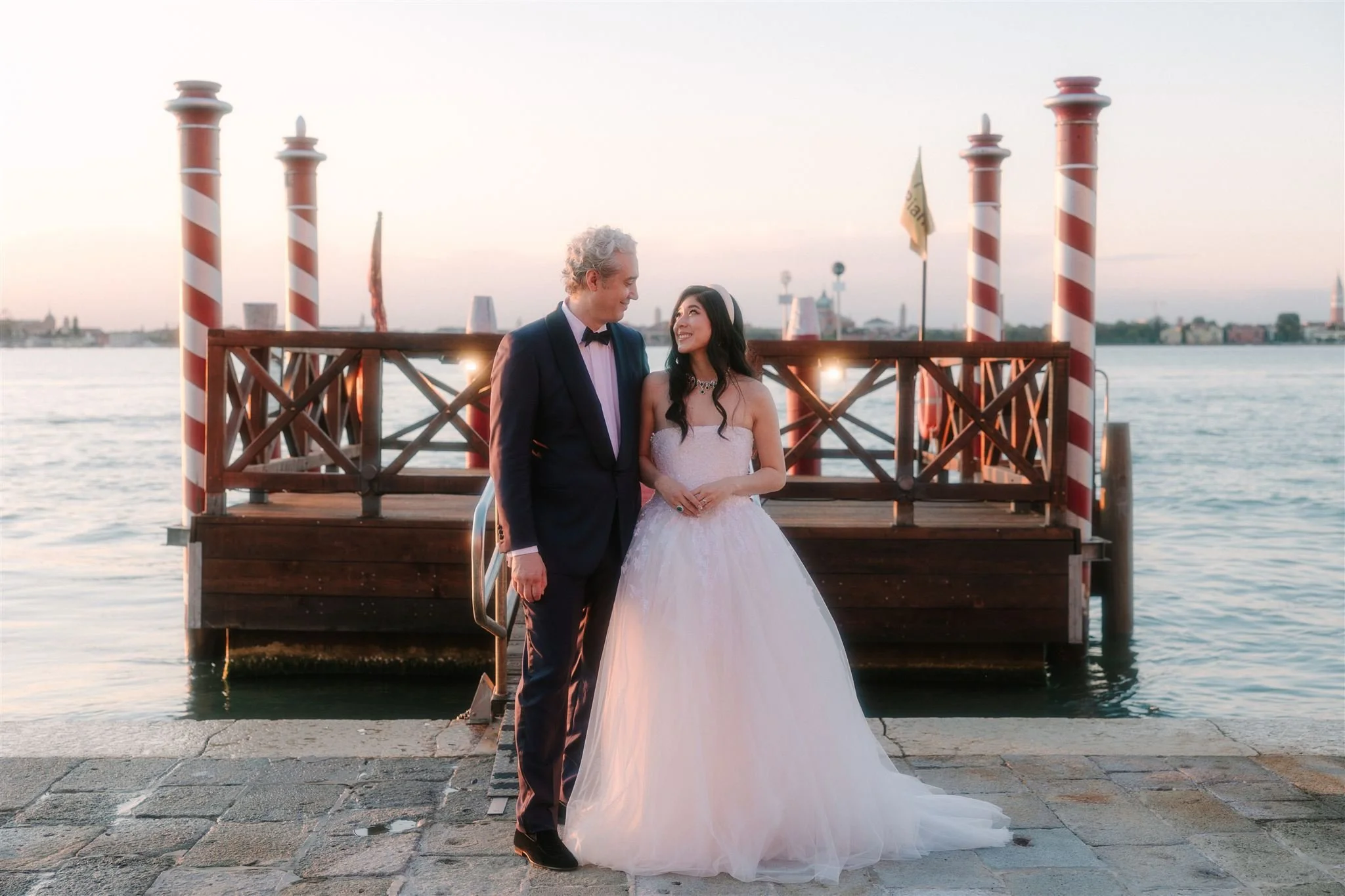 A bride and a groom in black tie looking at each other during sunset time near the edge of the water at their Wedding on San Clemente Island, Venice at the Kempinski Hotel, with the pier behind them and the Venetian little dock with traditional poles