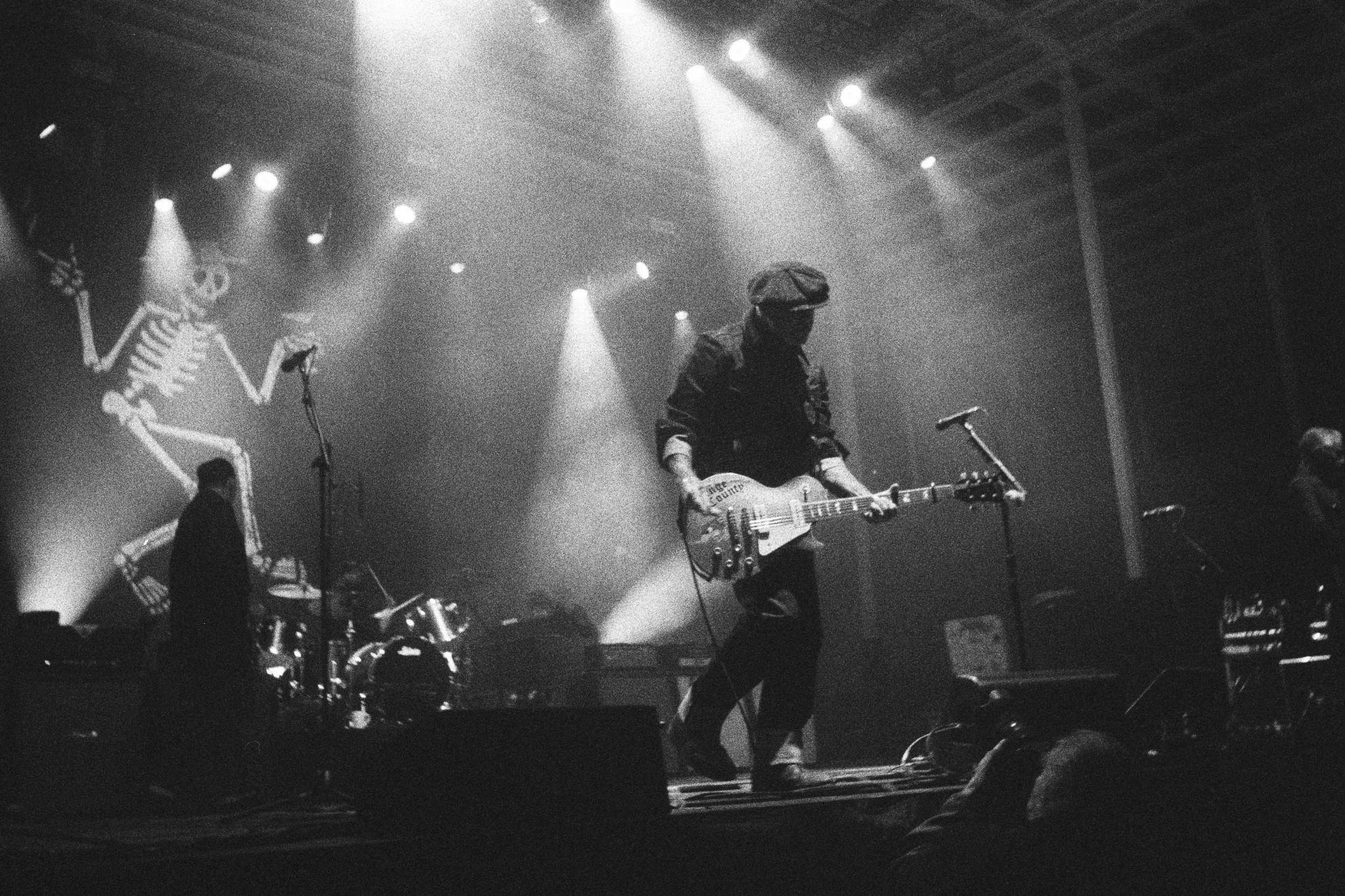 Black and white photo of a band performing on stage with a guitarist and singer Mike Ness in the foreground and drums in the background. Stage lights shine down on the performers, and a banner is visible on the wall behind them.