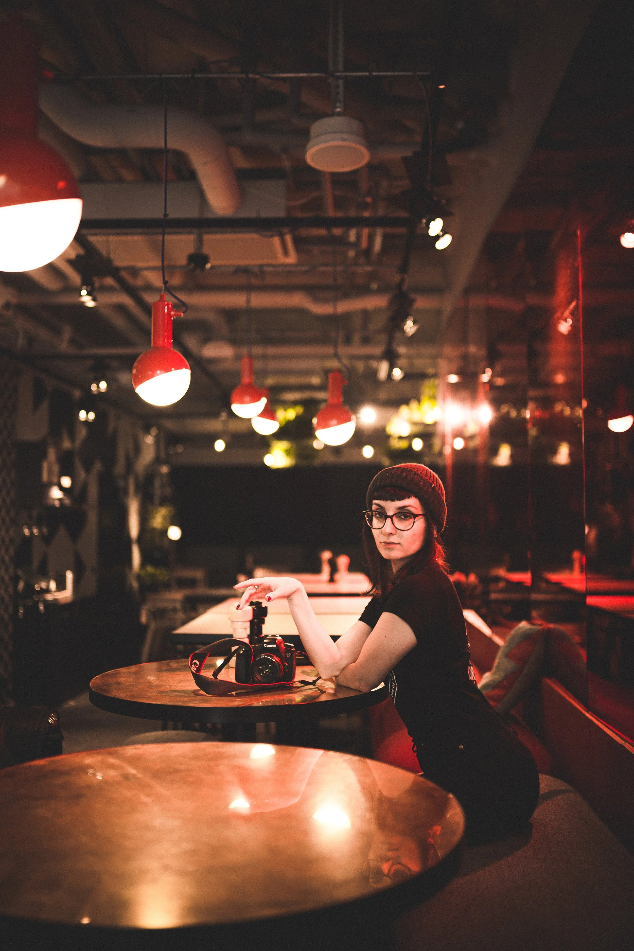 Young woman in a black beanie and glasses sitting at a table in a dimly lit cafe with red pendant lights, holding a camera.
