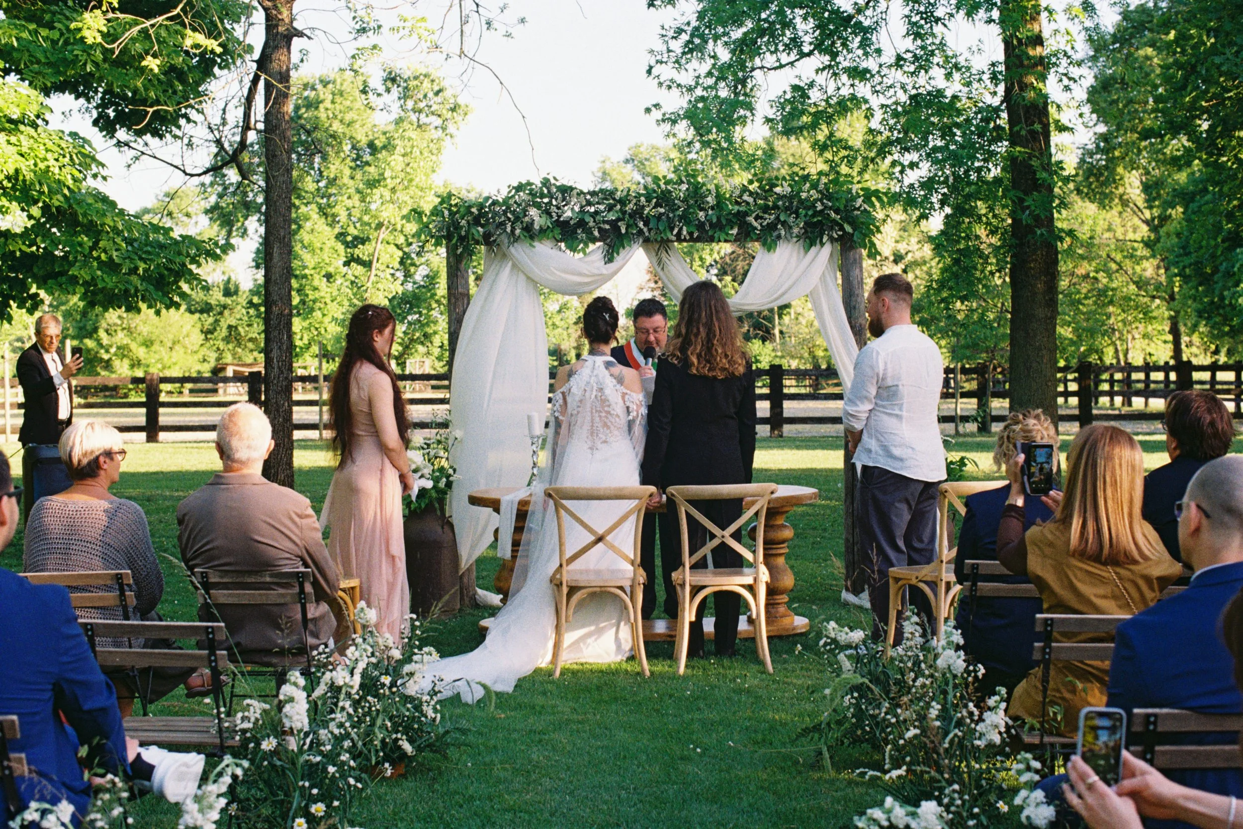 35mm film photo of outdoor wedding ceremony at mondelli stables taking place under a decorated arch with white drapes and green foliage, with guests seated and taking photos in a lush green park setting.