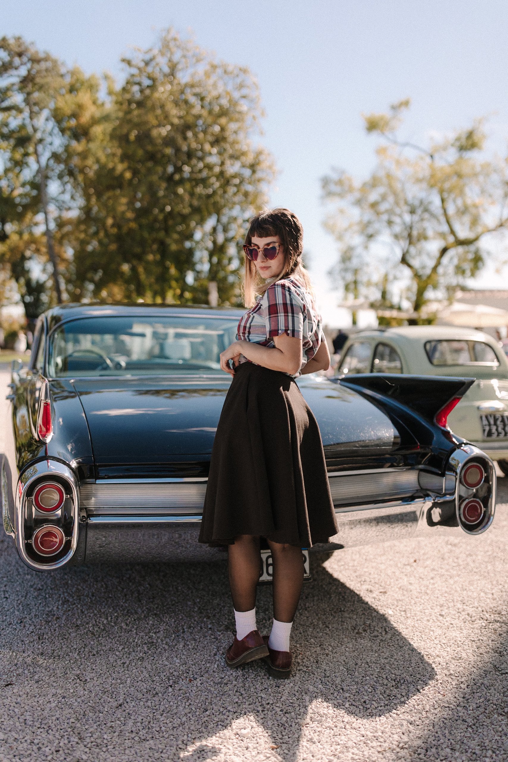 A woman wearing sunglasses, a plaid shirt, a black skirt, white socks, and brown shoes stands in front of a vintage black Cadillac at a car show during daytime.