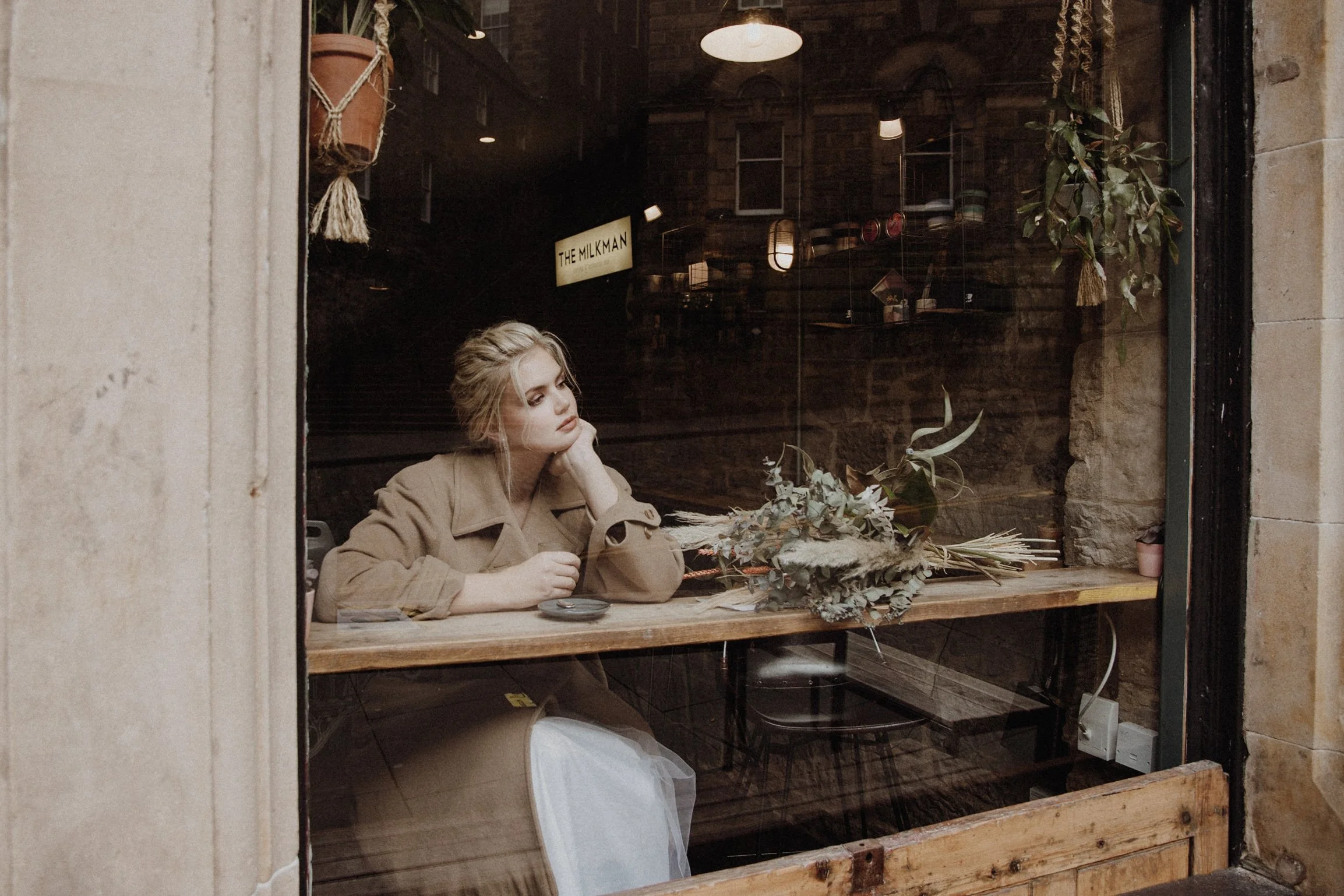Bridal model with a white wedding gown and a camel coat sitting on the window of Milkman cafe in Edinburgh holding a coffee mug and looking outside, with a dried flower bouquet next to her