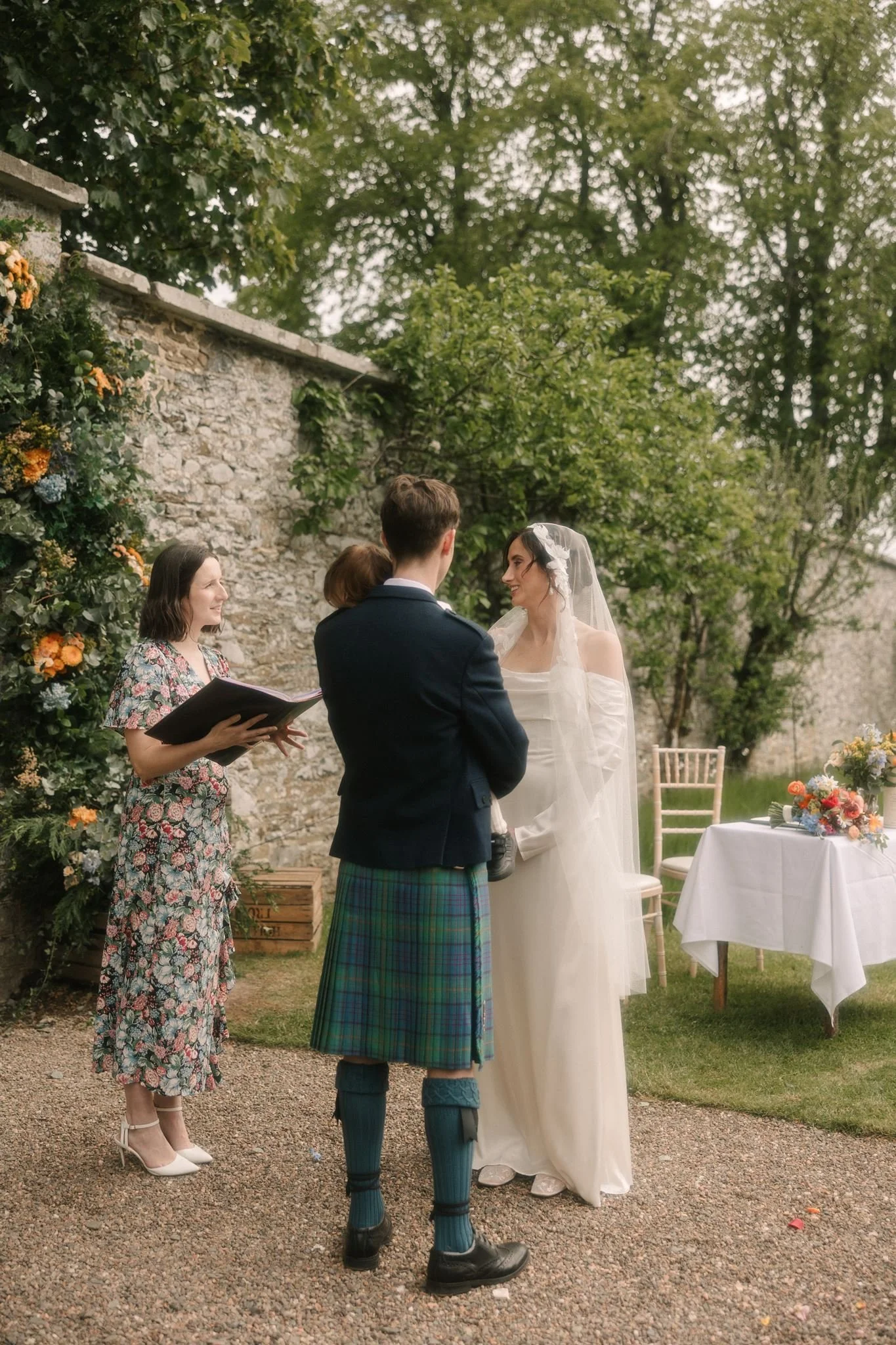 A wedding ceremony outdoors with a bride and groom facing each other, surrounded by friends and family, during daytime. The bride wears a wedding dress with veil, the groom is in a suit with kilt. A woman reads from a book, and there are flowers and chairs in the background.