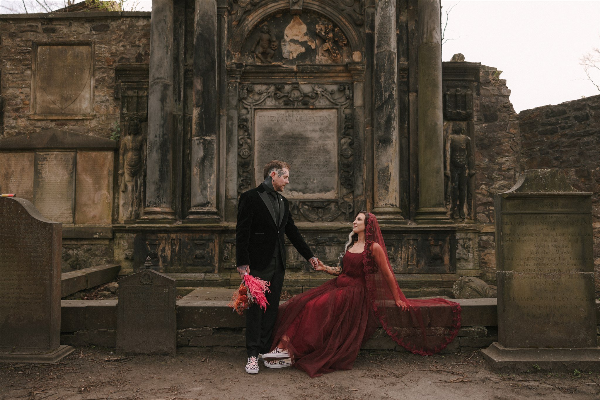 Alternative wedding couple with the bride dressed in a deep red gown and veil sitting in front of a tombstone in Edinburgh greyfriars Graveyard with the heavily tattooed groom standing next to her holding her hand and looking at each other