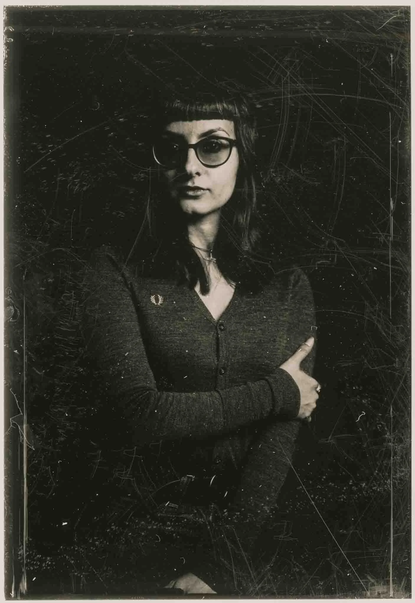 Black and white portrait of a woman with dark hair and bangs, wearing glasses, a dark top, and a necklace, with her arms crossed against a textured black background.