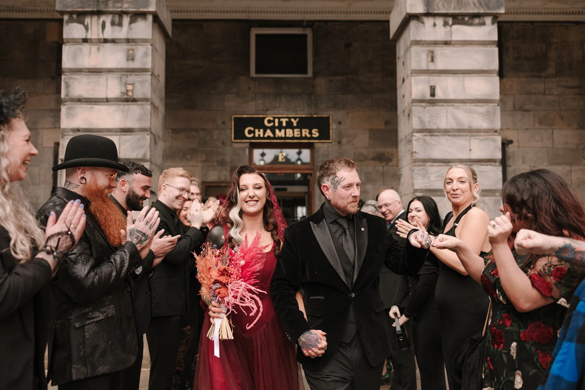 Alternative wedding couple with bride in a red wedding dress and heavily tattoed groom exiting Edinburgh City chambers after their ceremony, cheered by their guests