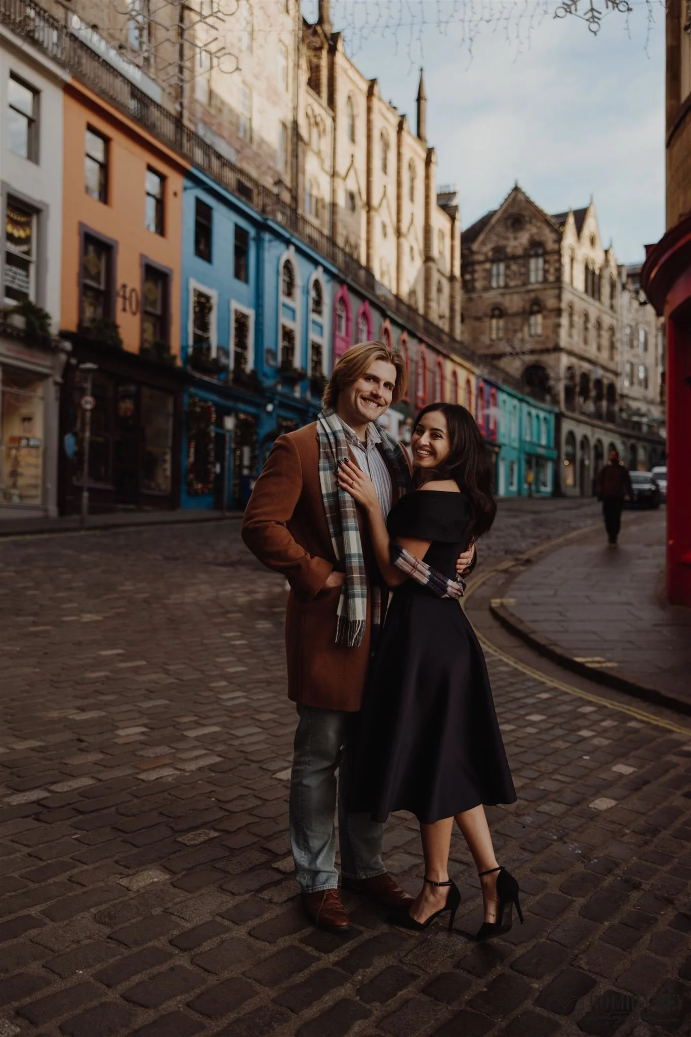 engaged Copule photographed hugging at the feet of Victoria Street in Edinburgh, with coloured buildings behind them and the curved cobbled street going uphill