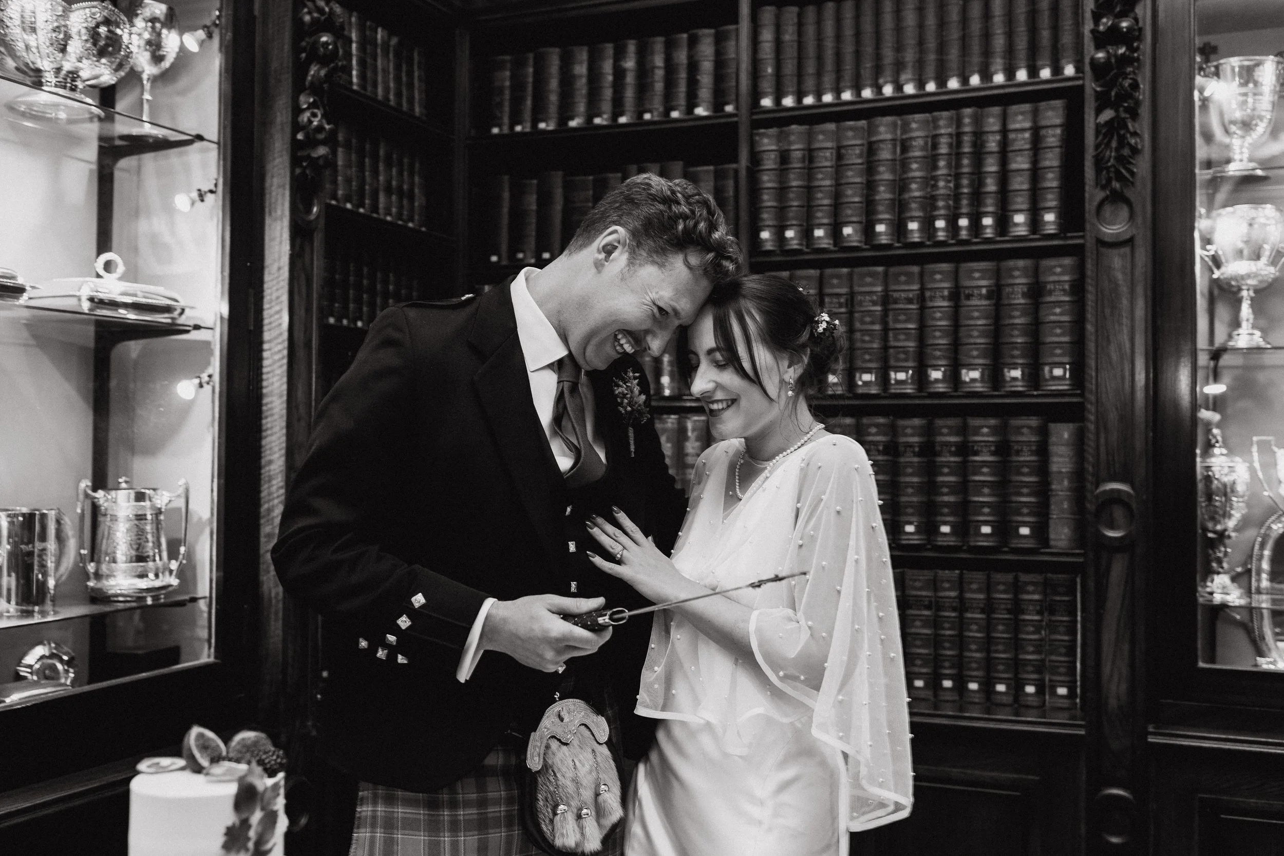 A wedding couple smiling and laughing inside a room with dark wooden bookshelves, glass display cases with trophies, and a cake with figs and strawberries on top.