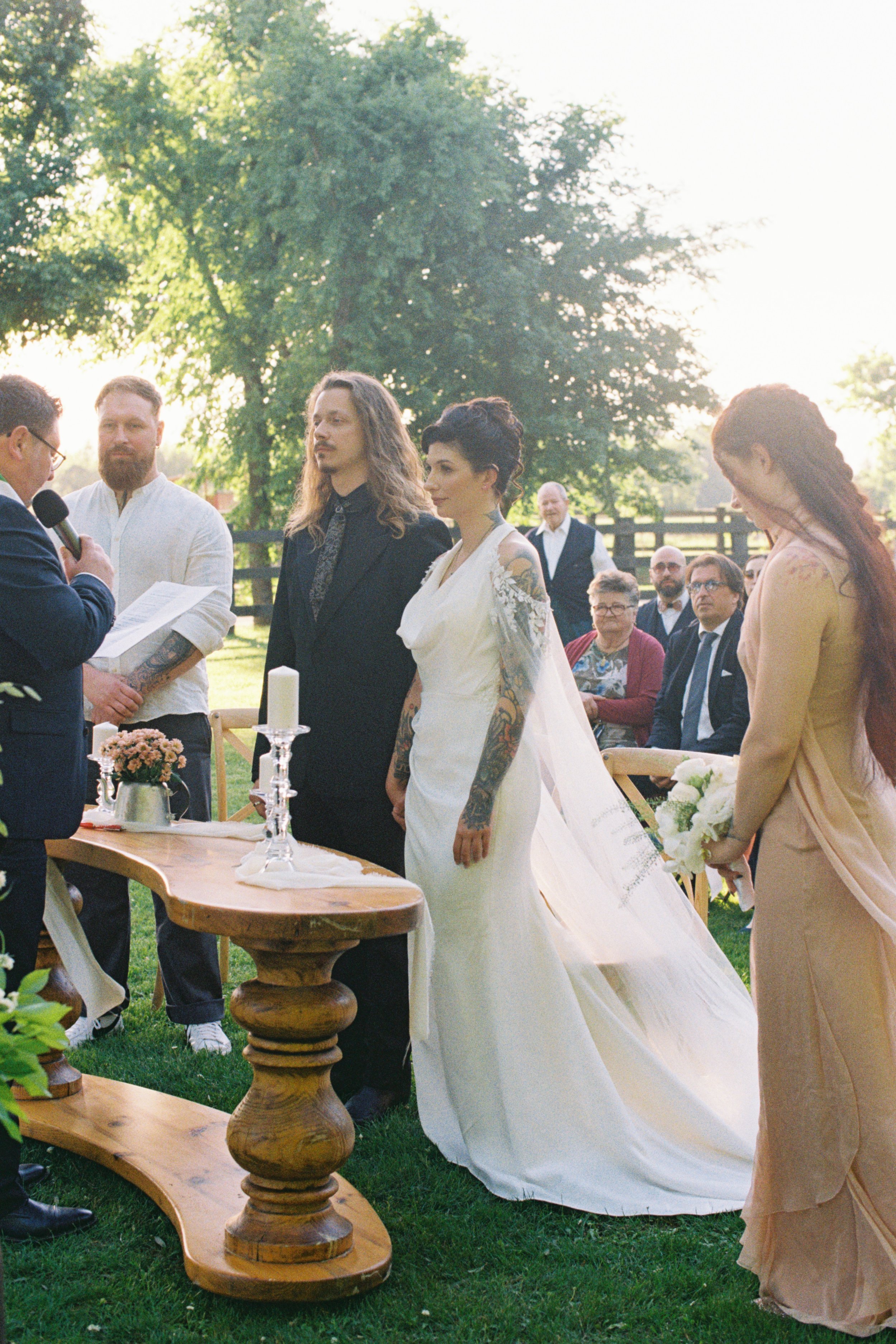 A wedding ceremony outdoors with a bride in a white wedding dress and tattoos standing in front of an officiant. Groomsmen and guests are seated and standing, with a large tree and fence in the background, during the daytime.