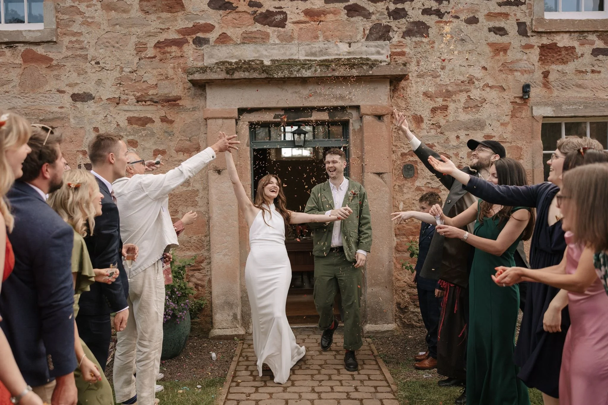 People celebrating a wedding outside a stone building, with a bride and groom smiling and surrounded by wedding guests.