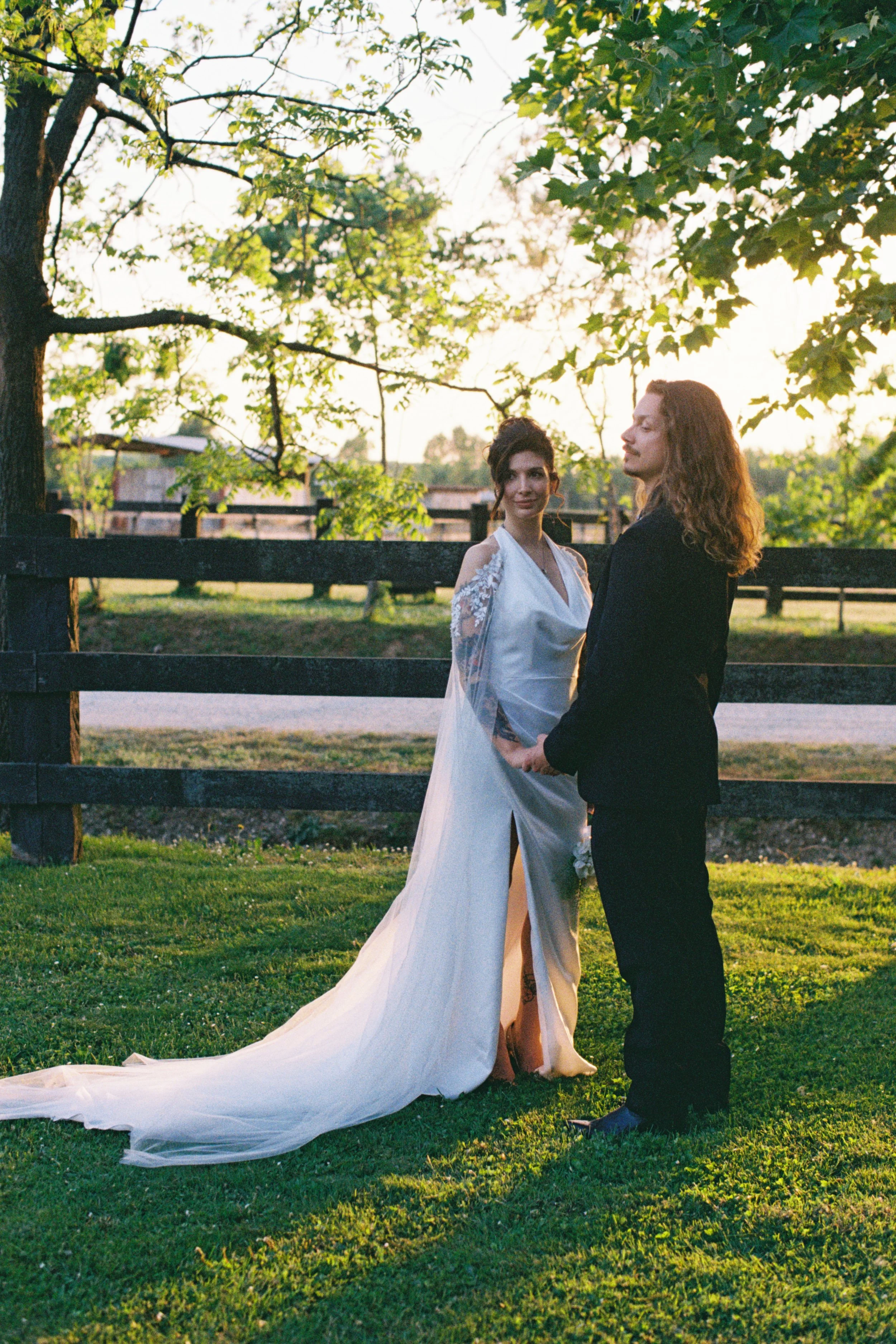 A bride and groom holding hands outdoors near a black wooden fence, with trees and a sunset sky in the background.