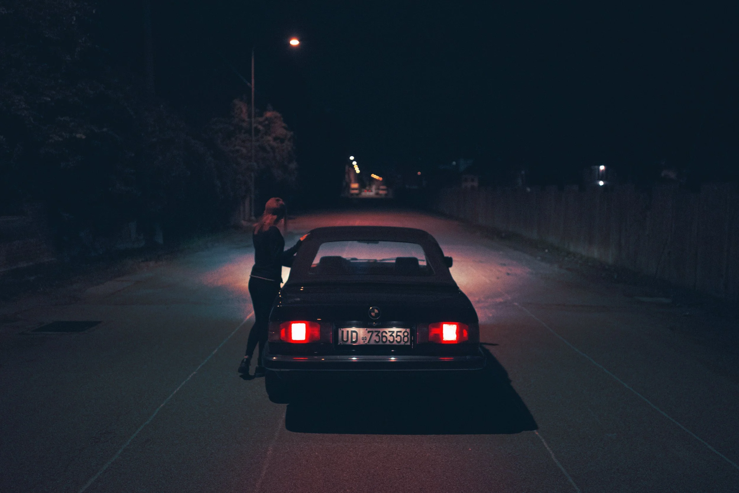 A woman standing next to a black convertible car at night on a deserted street, with streetlights in the distance.