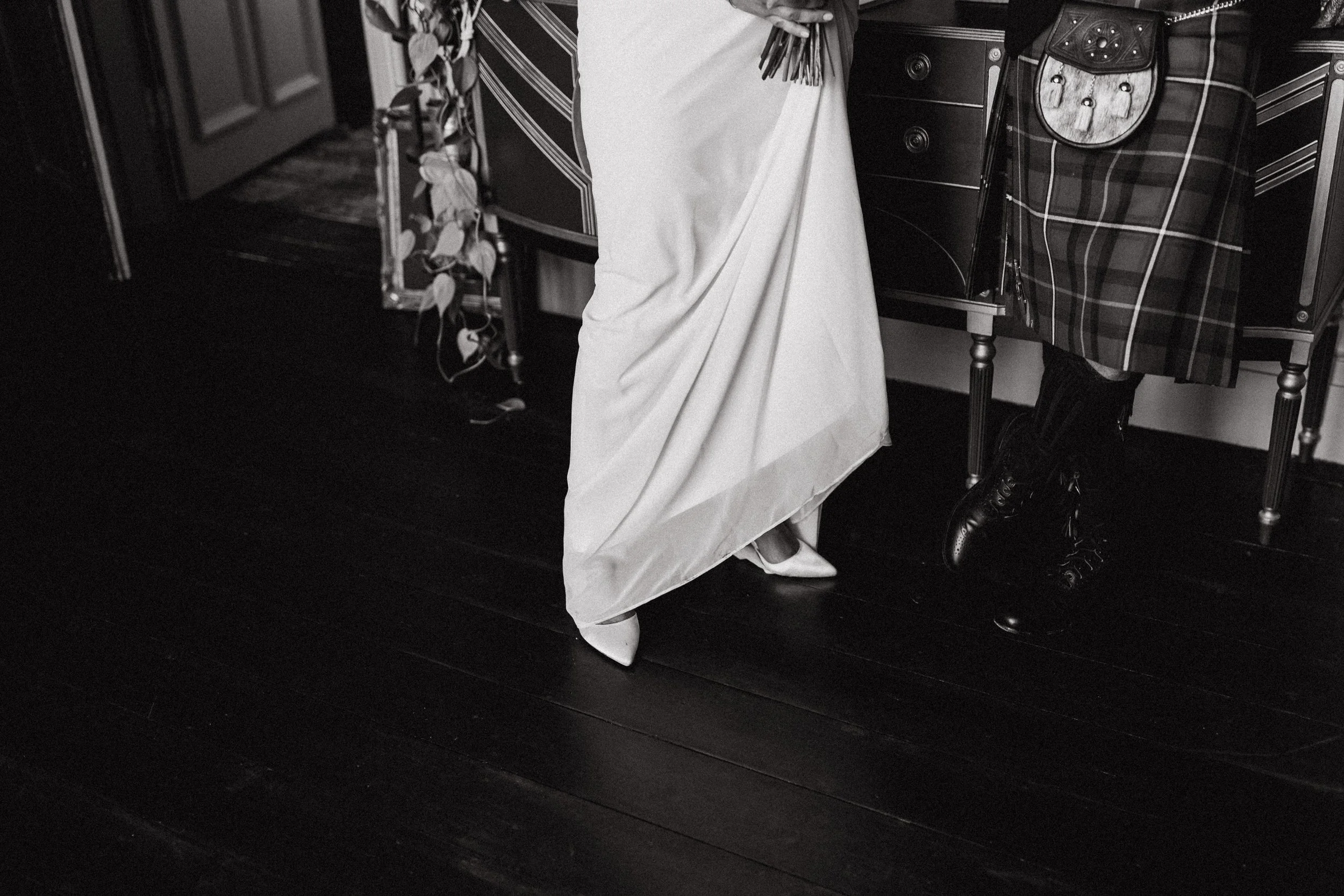 A person in a wedding dress and high heels standing next to a person in a tartan skirt with combat boots, seated at a table in a room with wooden flooring.