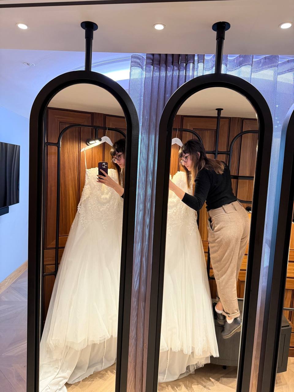 A woman taking a mirror selfie with a wedding dress, with a wooden panel background and a gray curtain, using two full-length mirrors.