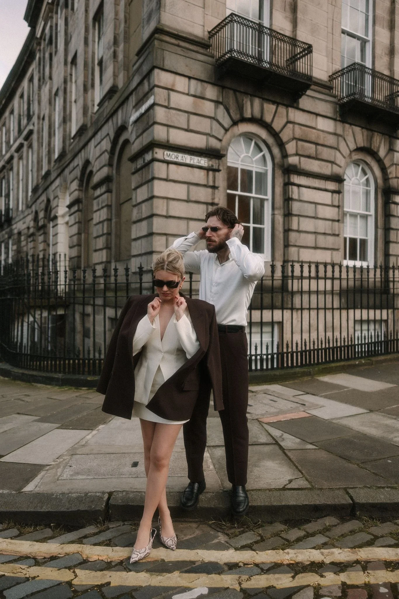 A man and woman walking on a city sidewalk in front of a historic building with arched windows and a sign that reads 'Moray Place'. The woman is wearing a white blazer, beige high heels, sunglasses, and a dark brown blazer over her shoulders. The man is dressed in a white shirt and dark trousers, also wearing sunglasses, and covering his ears with his hands.