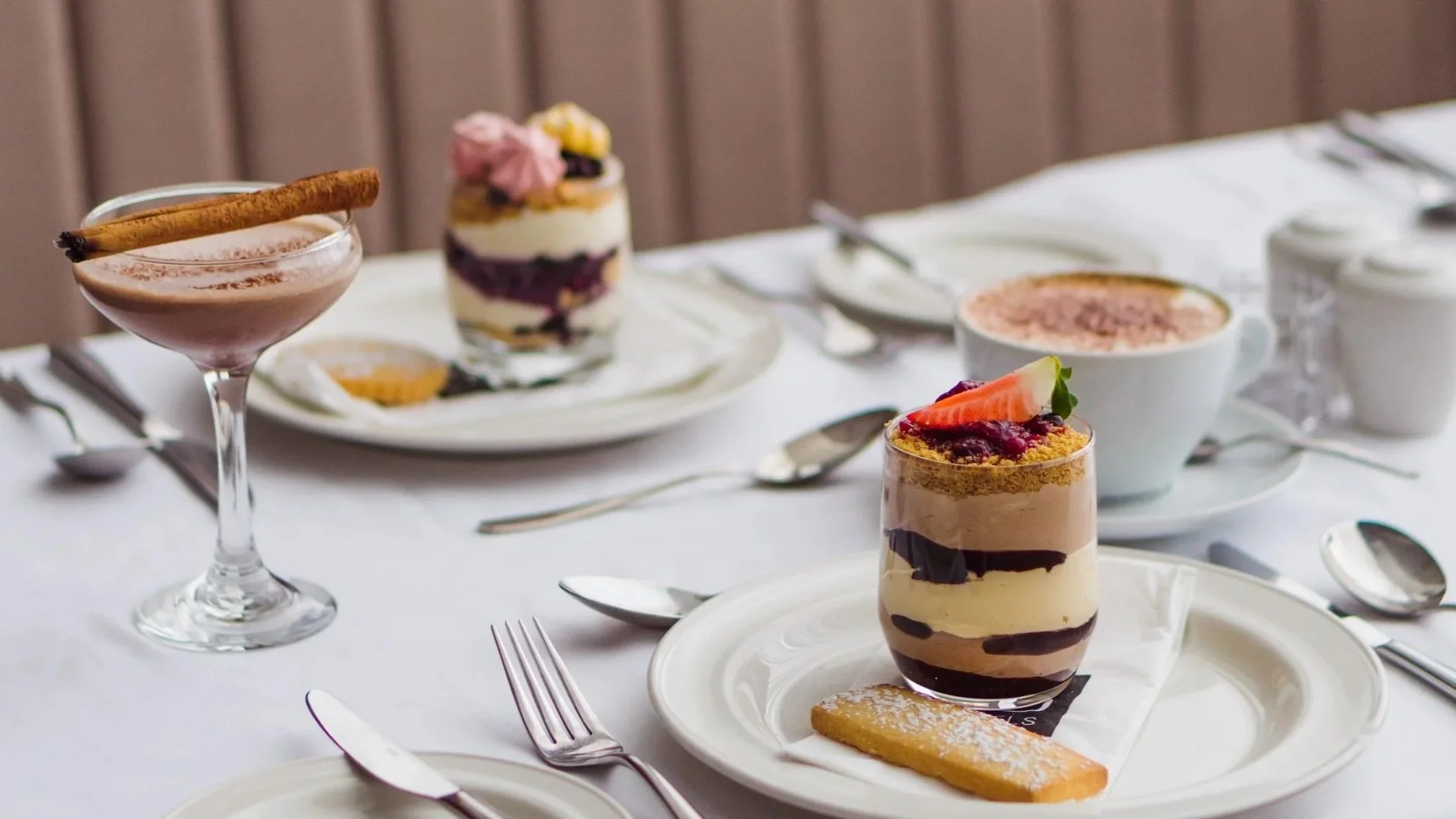 Table with desserts and drinks, including a parfait, cappuccino, and cocktail with cinnamon stick.