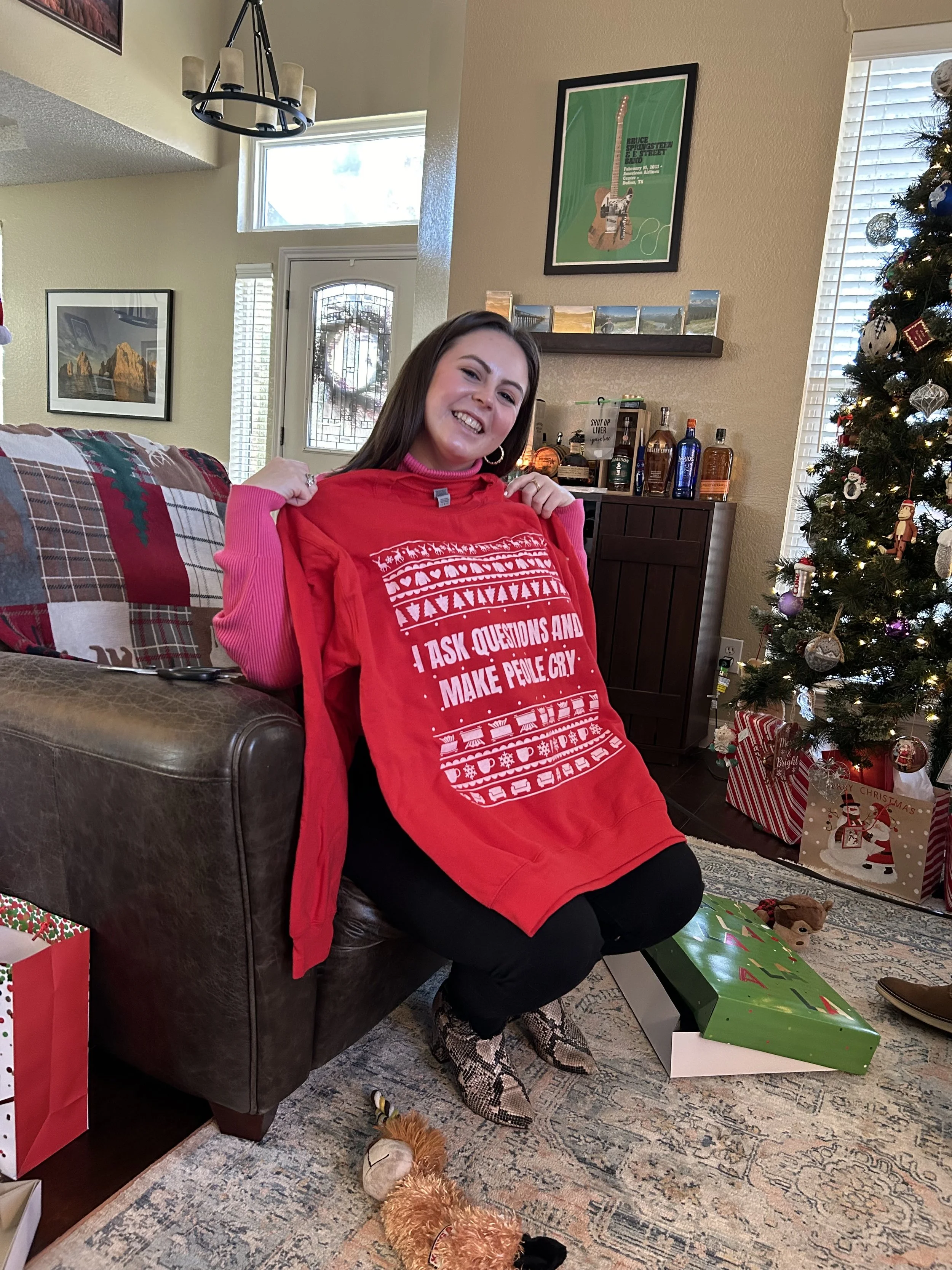 A woman smiling while holding up a red holiday sweater in a cozy living room with a Christmas tree and wrapped gifts, illustrating the joyful and stressful parts of holiday perfectionism and people pleasing.