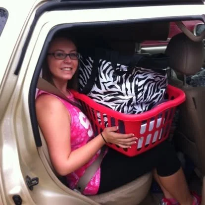 Young woman sitting in a packed car with a laundry basket and belongings while leaving for college, illustrating the planning and mental load involved in back-to-school transitions.