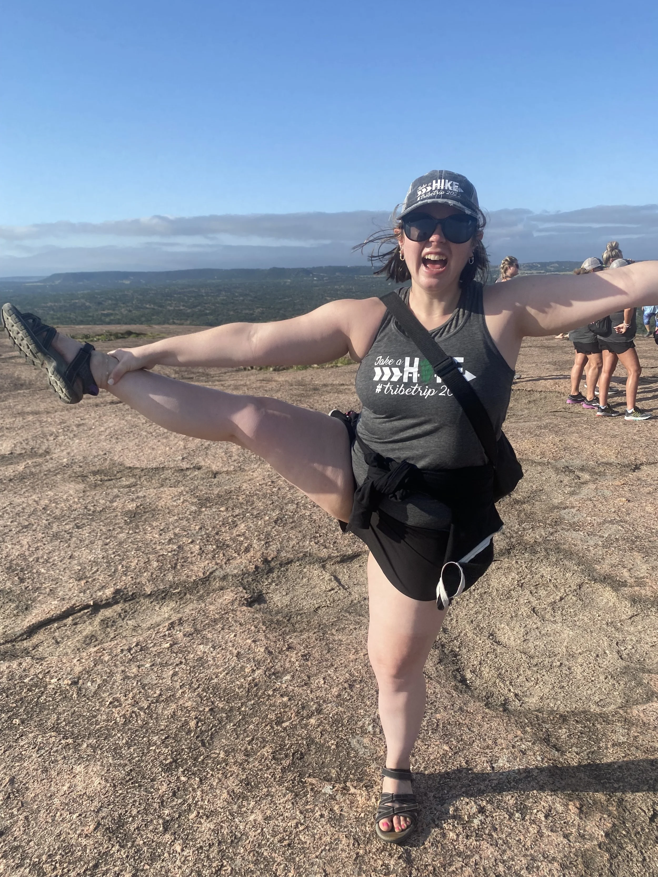 A woman smiling with arms open and leg lifted while standing on a rocky overlook, embodying joy, freedom, and confidence after leaving an emotionally unequal relationship and healing from emotional overfunctioning.