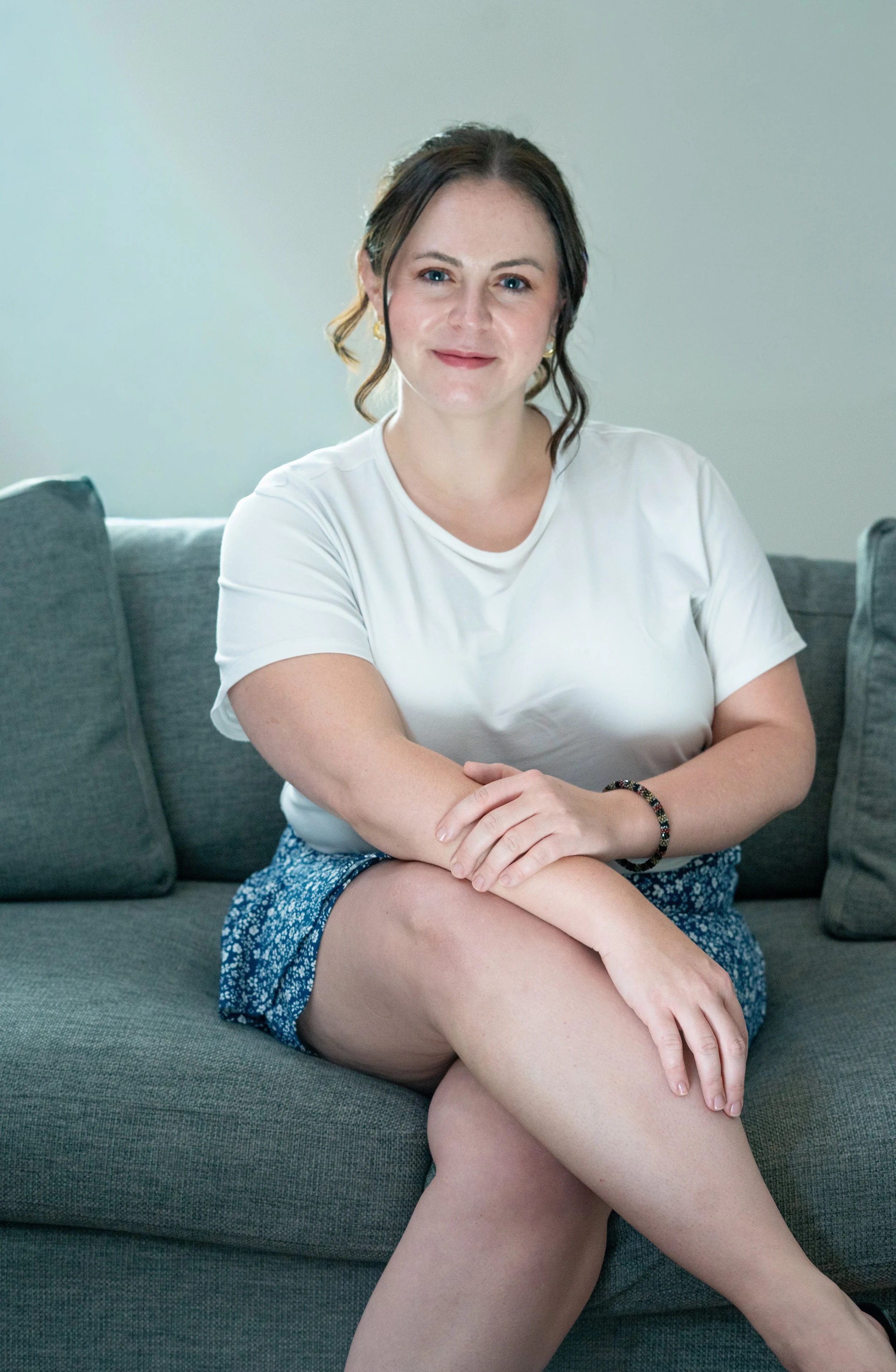 A woman with brown hair and fair skin sitting on a gray couch, wearing a white t-shirt and a blue patterned skirt, with legs crossed and hands resting on her knee, looking at the camera with a slight smile.