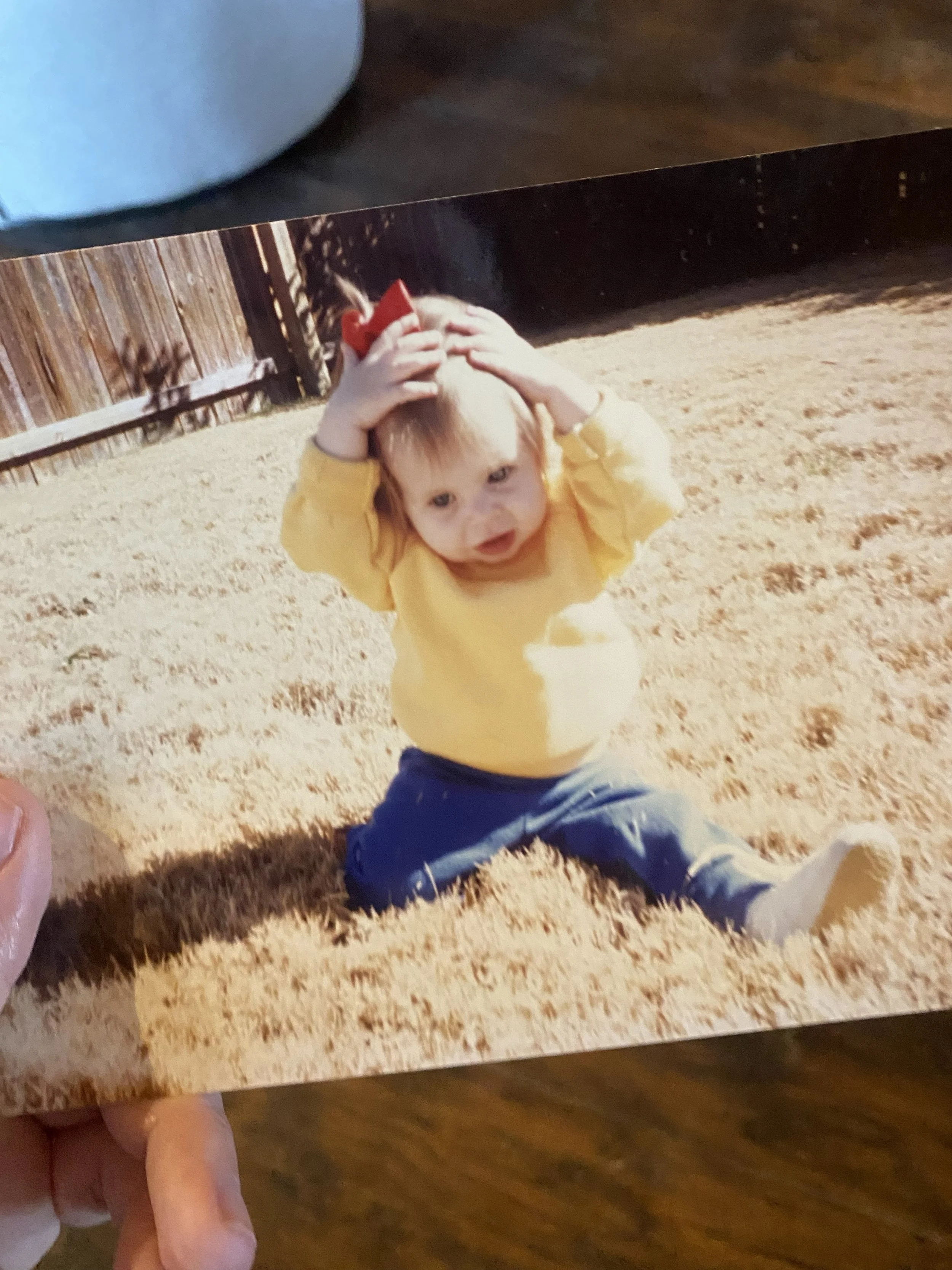 Child sitting outside in a yellow shirt with a red bow in her hair, holding both hands on top of her head.