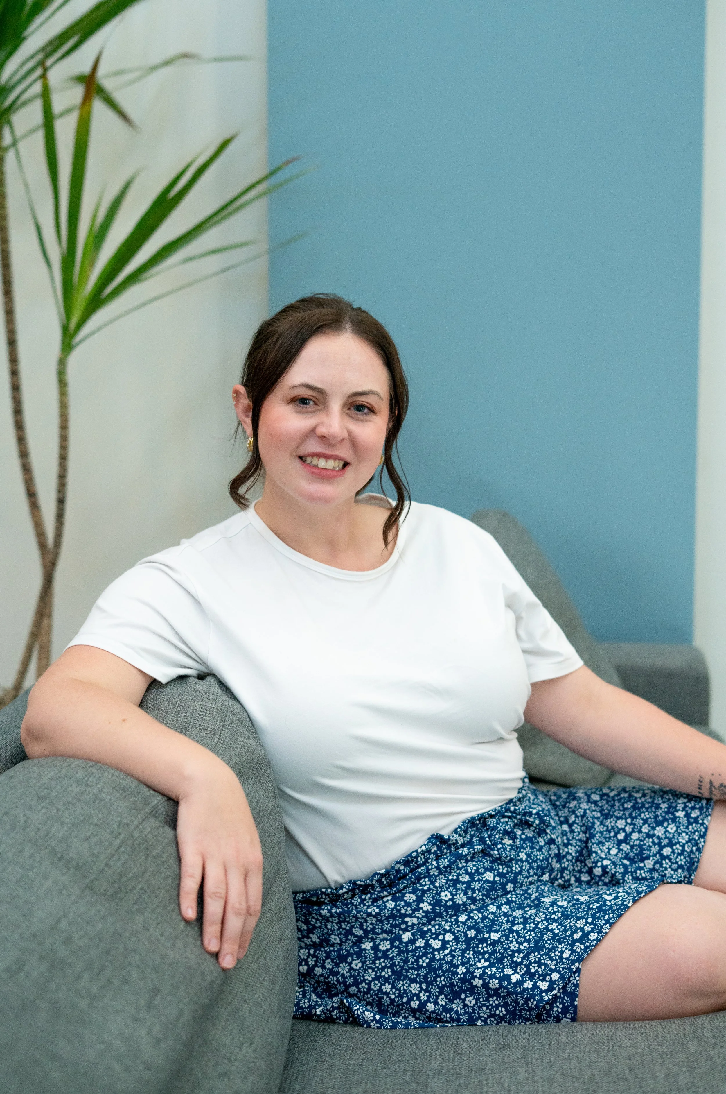 A woman with dark brown hair, wearing a white t-shirt and blue patterned shorts, sitting on a gray couch in a room with light blue and cream-colored walls and a tall green plant.