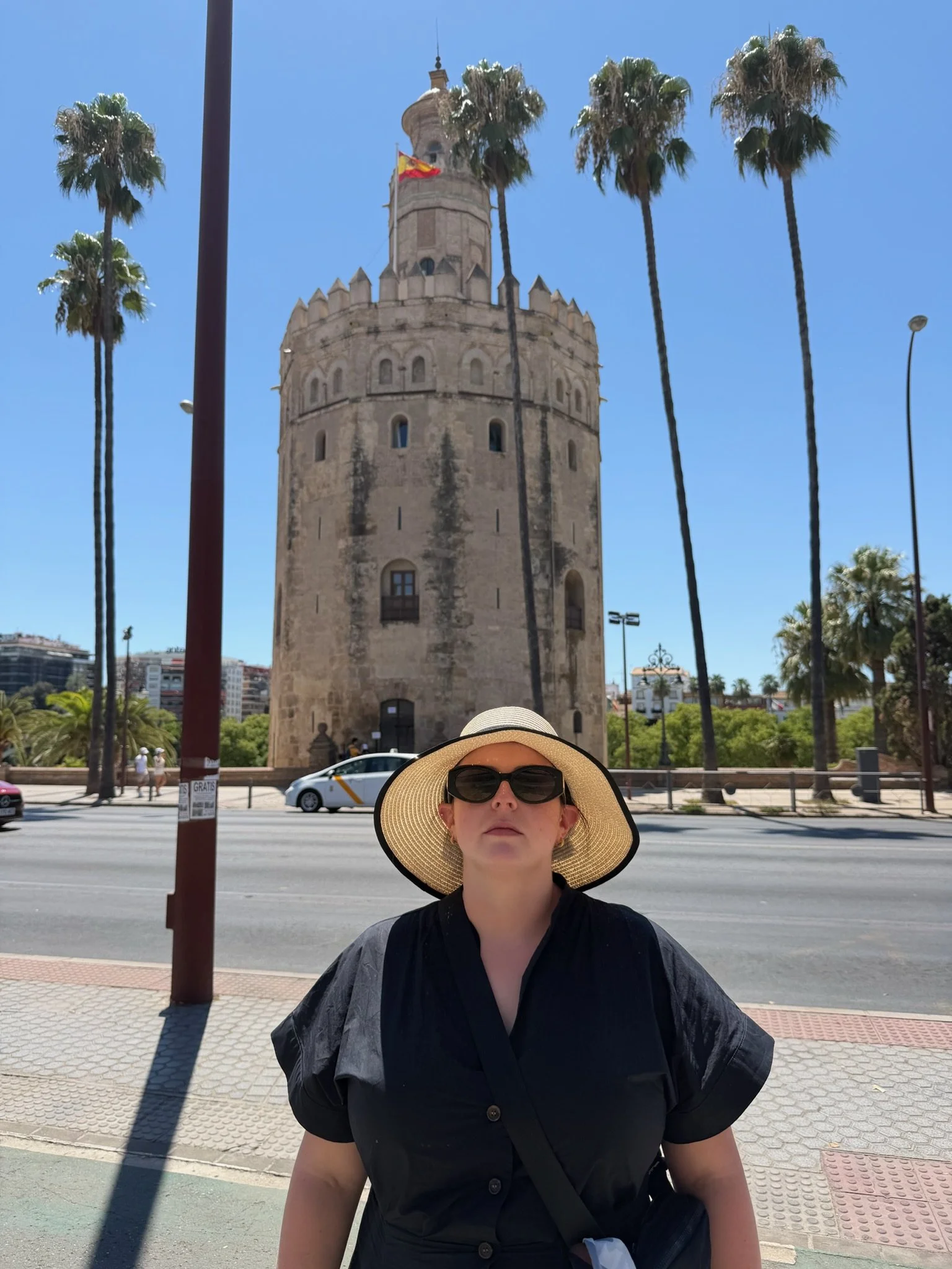 Therapist Aubrey Richardson wearing a sun hat and sunglasses while traveling in Seville, Spain, with the Torre del Oro and palm trees behind her.