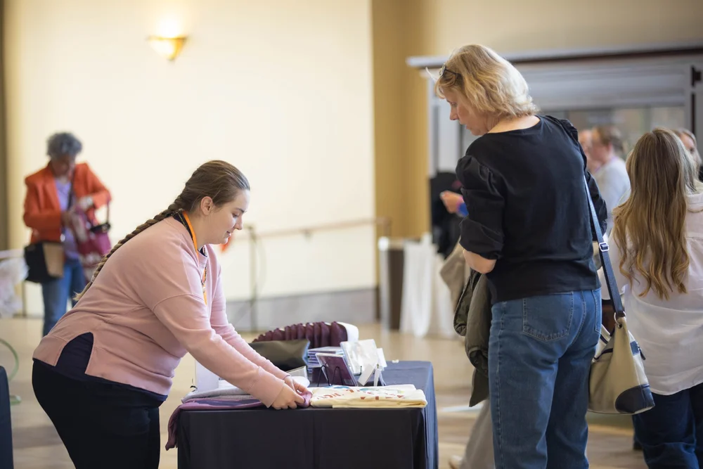 A volunteer in a MORE lanyard leans across a display table to help a guest.