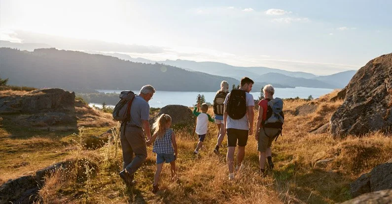 Group of people hiking on a grassy trail beside rocks with a lake and hills in the background during daytime.
