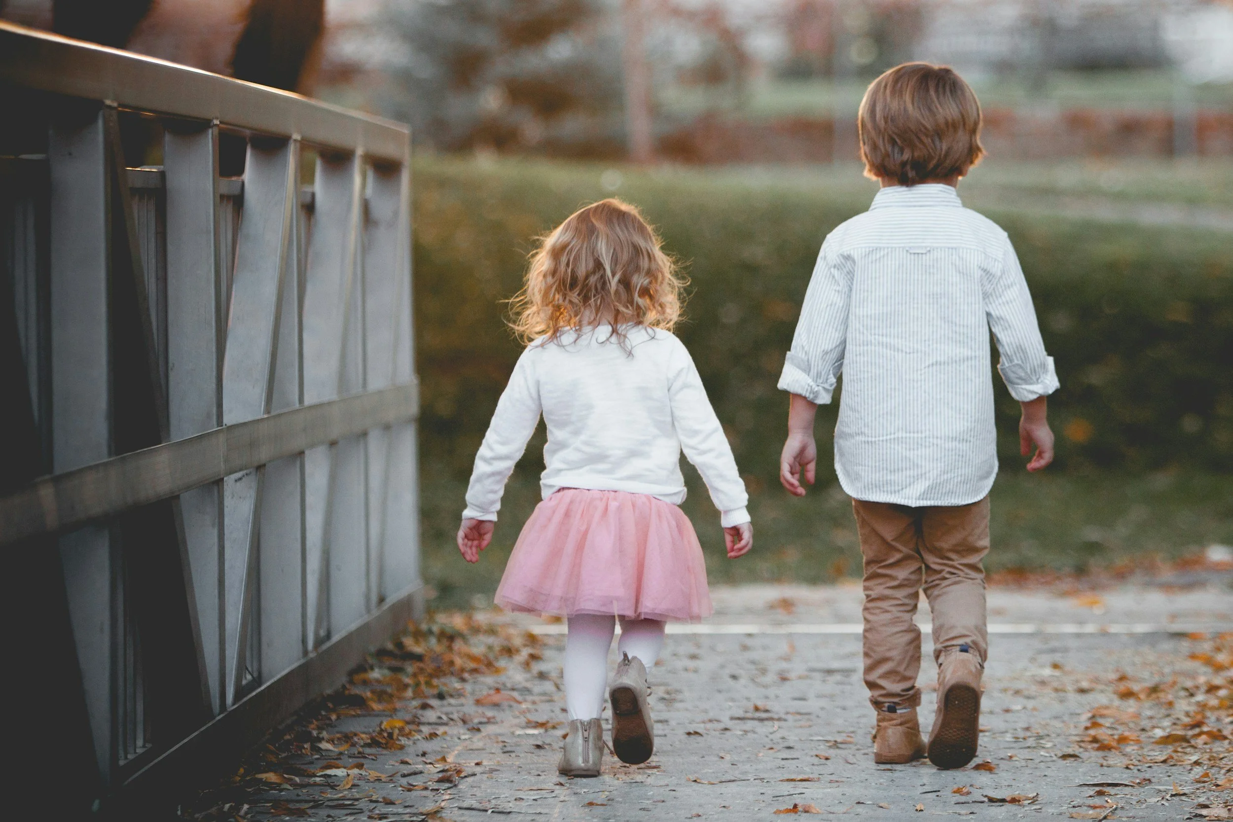 Two children, a girl and a boy, walking away on a park pathway during autumn, with falling leaves and trees in the background.