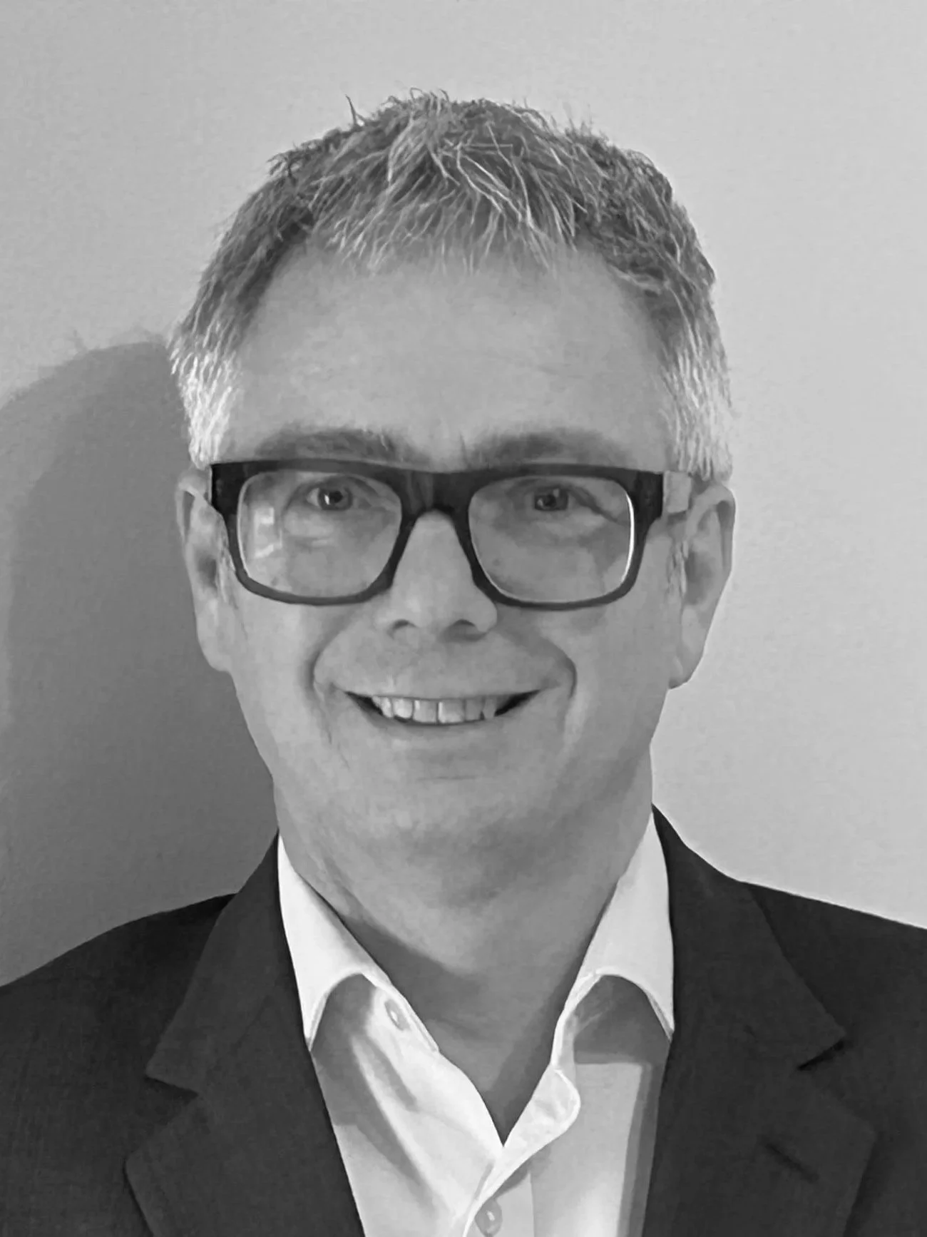 Black and white portrait of a man with glasses, short gray hair, dressed in a suit and white shirt, smiling at the camera.
