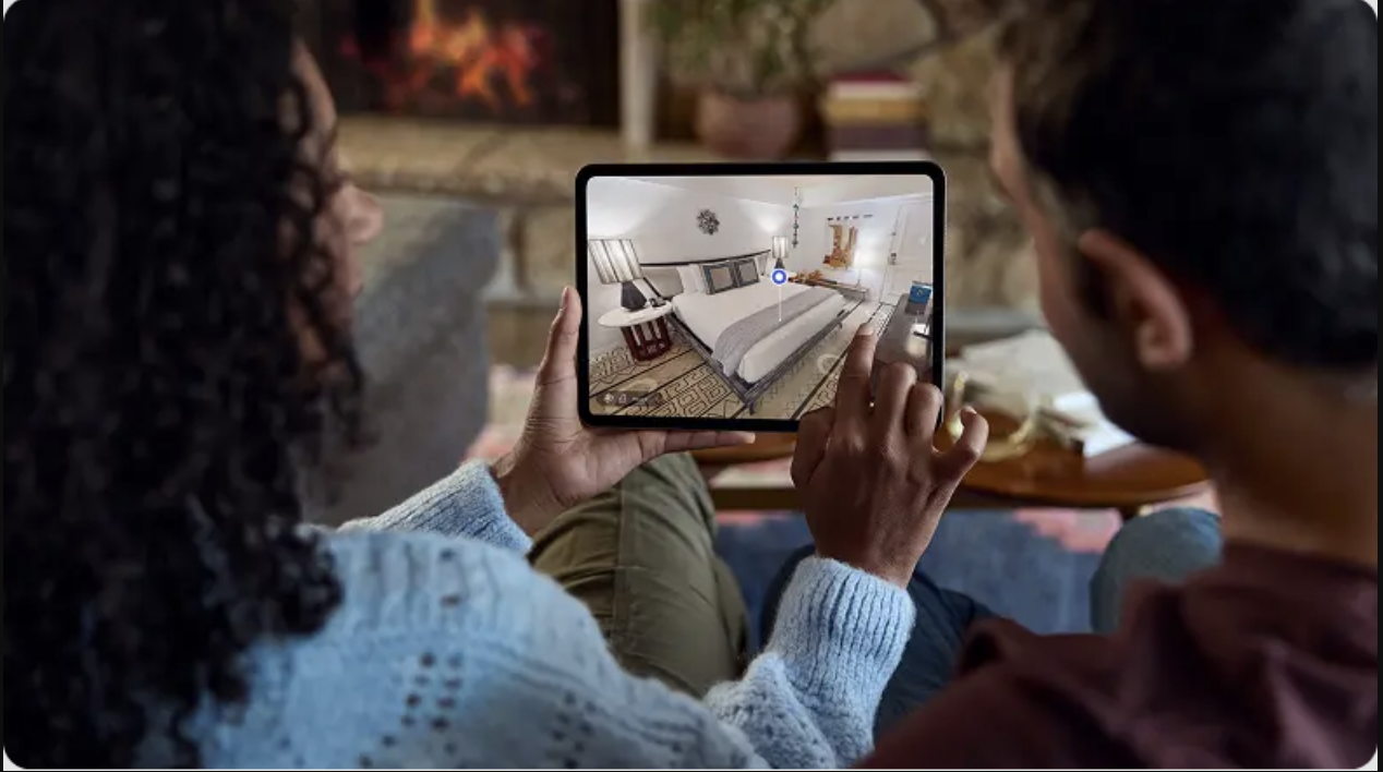 Two people sitting on a couch looking at a tablet showing a 3D view of a bedroom.