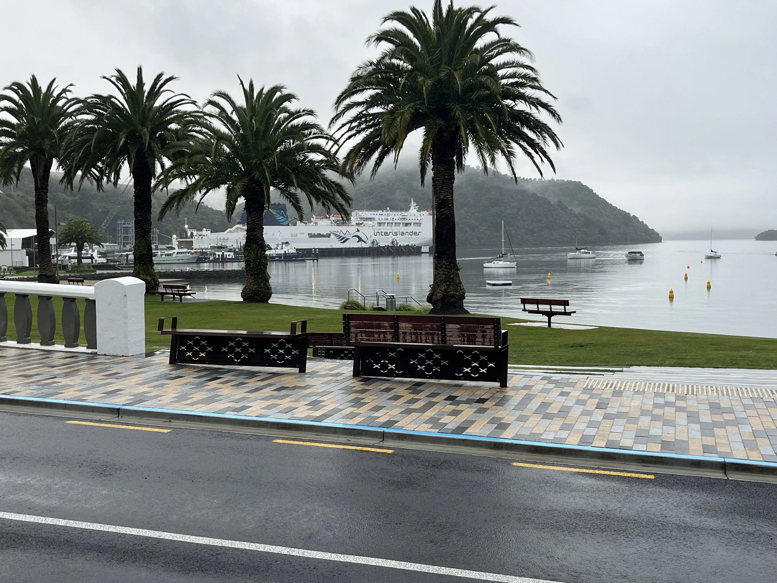 View over road and out onto water at Picton shwing bench seating with whetu design by Keni-Duke Hetet architectural designer, also some palms in the background and the interislander ferry