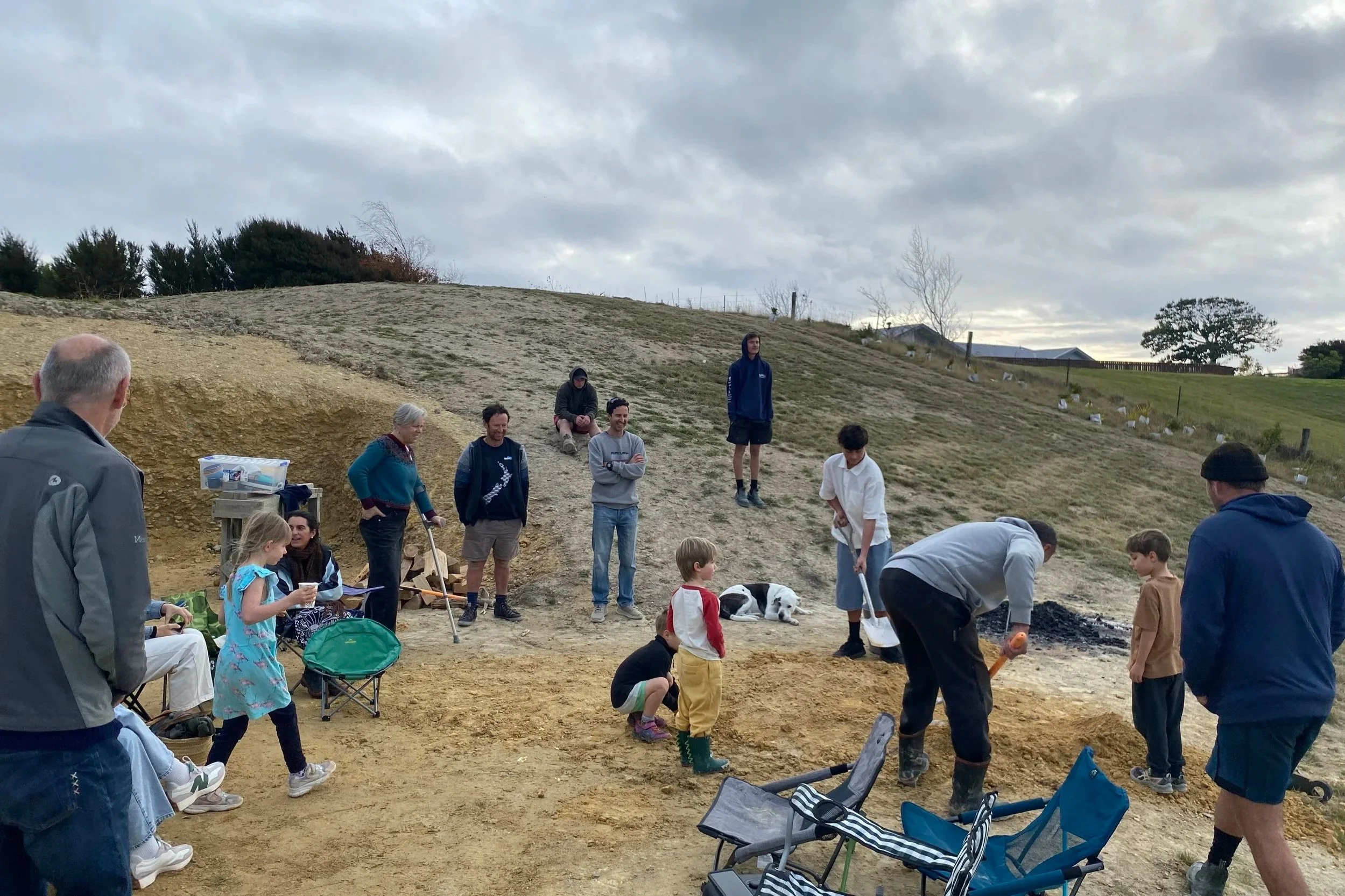 Group of people gathered outdoors on a hillside, some standing and some sitting, during cloudy weather. Children and adults are engaged in activities around the hangi site, with trees and a cloudy sky in the background.