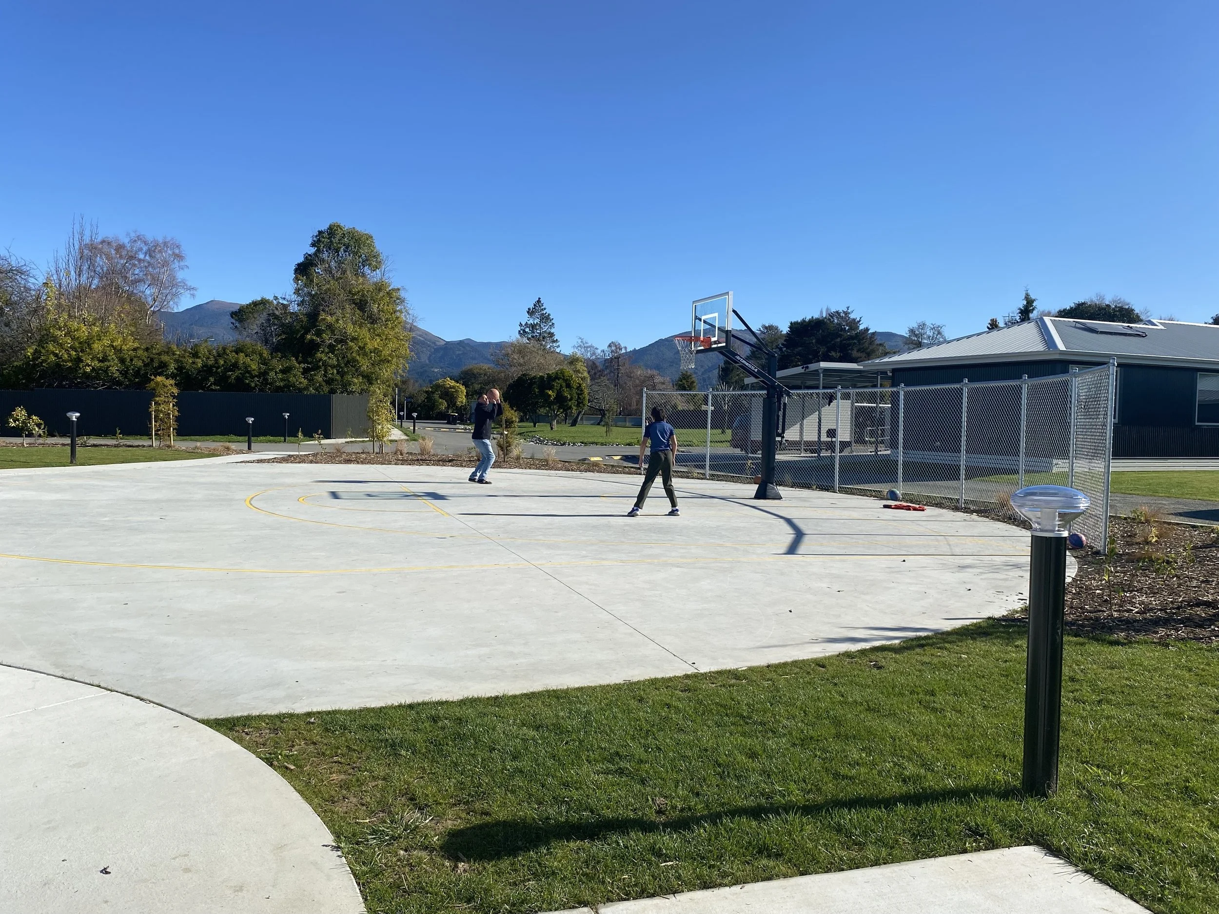 Basketball court sits adjacent to communal building at Te Awhina Marae papakainga, two people playing basketball on a bellbird blue day