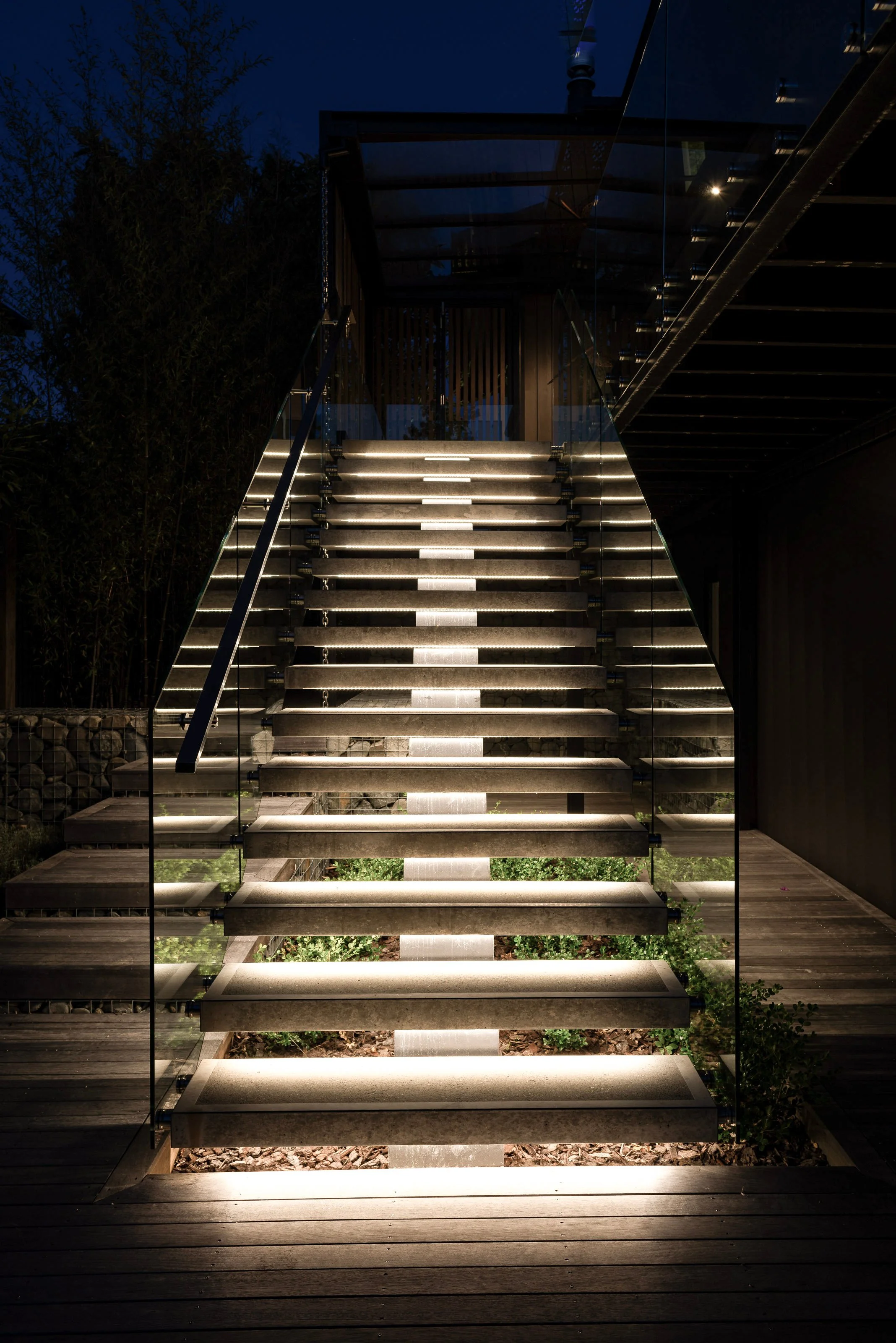 Stairs at Motueka Home lit up by under stair white lighting at night with a glass balustrade