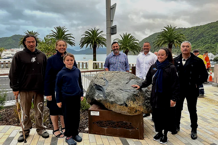 Group of people associated with the redevelopment of London Quay including architectural designer Keni-Duke Hetet standing around a large carved stone boulder on a pedestal outdoors, with palm trees, a body of water, and hills in the background.