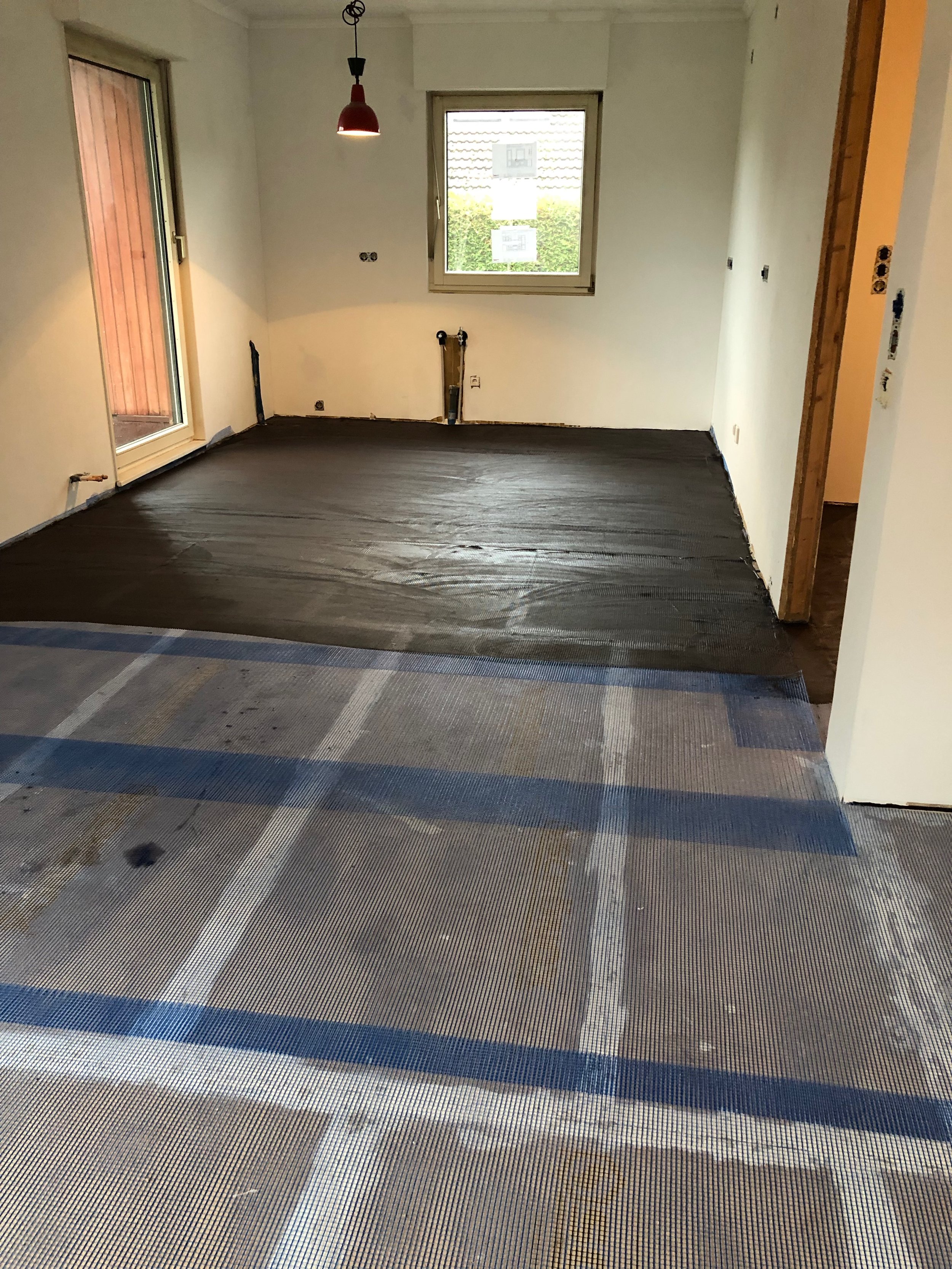 Ein Raum im Bau mit unbedecktem Boden, im Hintergrund ein Fenster, eine Tür führt zu einem Balkon, die Wände sind weiß und unvollständig, es sind mehrere elektrische Anschlüsse sichtbar, eine rote Deckenleuchte hängt von der Decke.