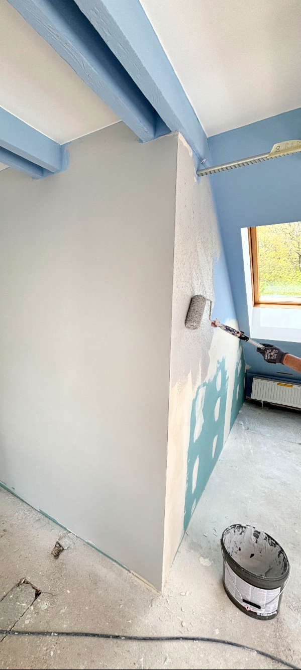 Maler streicht eine weiße Wand in einem Raum mit blauen Deckenbalken, neben einem Fenster, das Tageslicht einfällt.