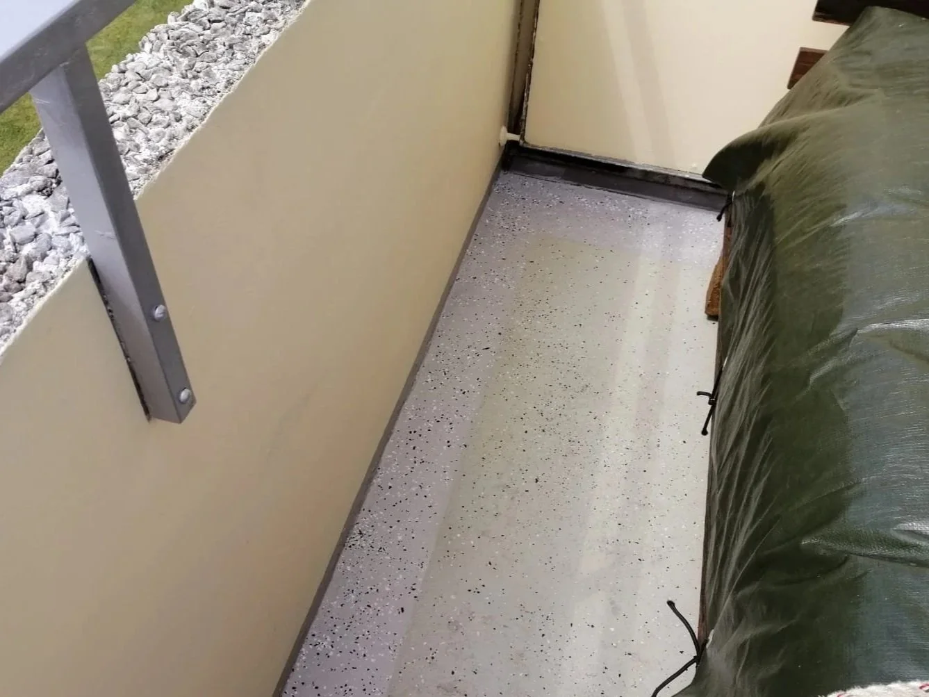 Balkon mit beige Wände, Sichtschutz aus grünem Plastik, heller Boden mit schwarzen Flecken, teilweise verdeckt durch eine Plane.