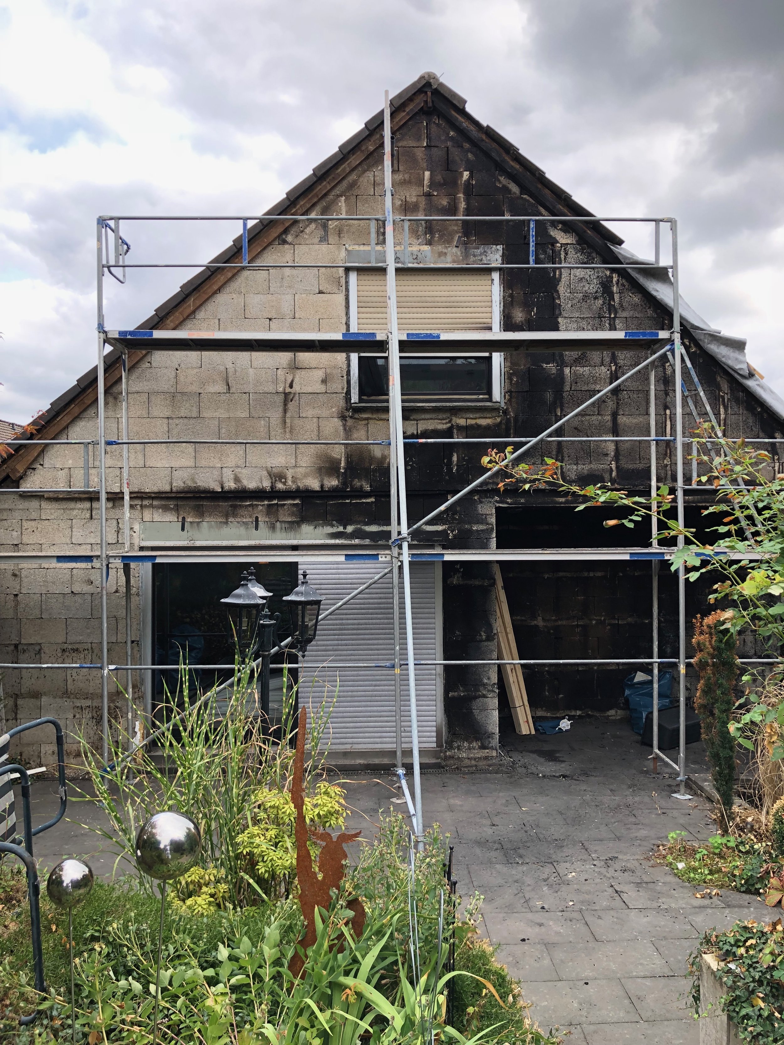 Haus im Umbau mit Gerüst, einige Fenster sind schwarz verkohlt, im Vorgarten sind Pflanzen, eine Laterne und dekorative Metallskulpturen.