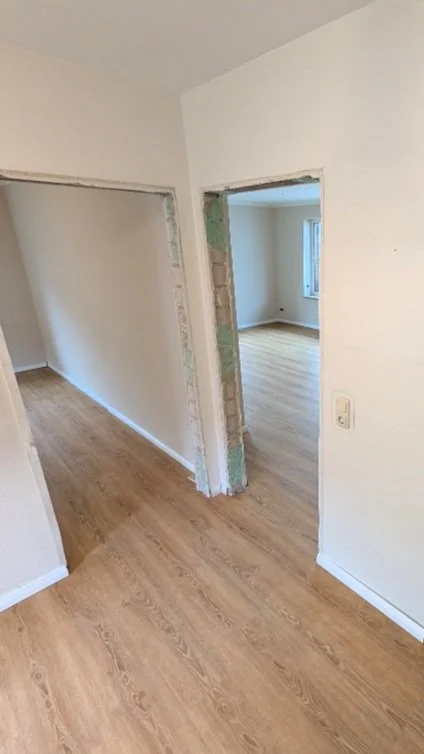 Innenbereich eines Hauses, derzeit im Renovierungsprozess, mit weißen Wänden, Holzfußboden und offenen Türrahmen, die in weitere Räume führen.