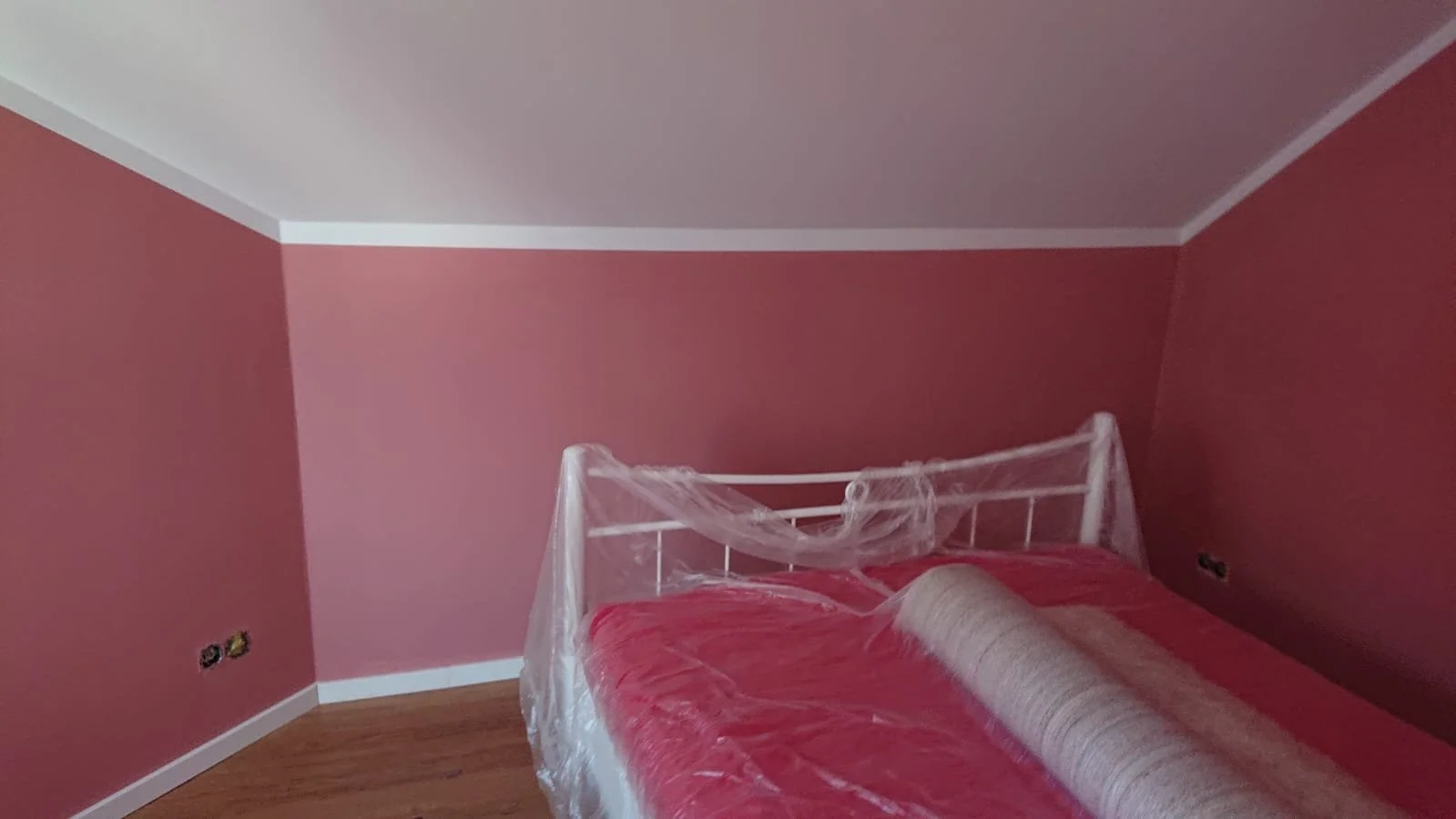 Ein Schlafzimmer mit roter Wandfarbe, einem weißen Bettrahmen, das Bett ist noch eingepackt mit Plastikfolie und einer Schutzfolie auf der Matratze.