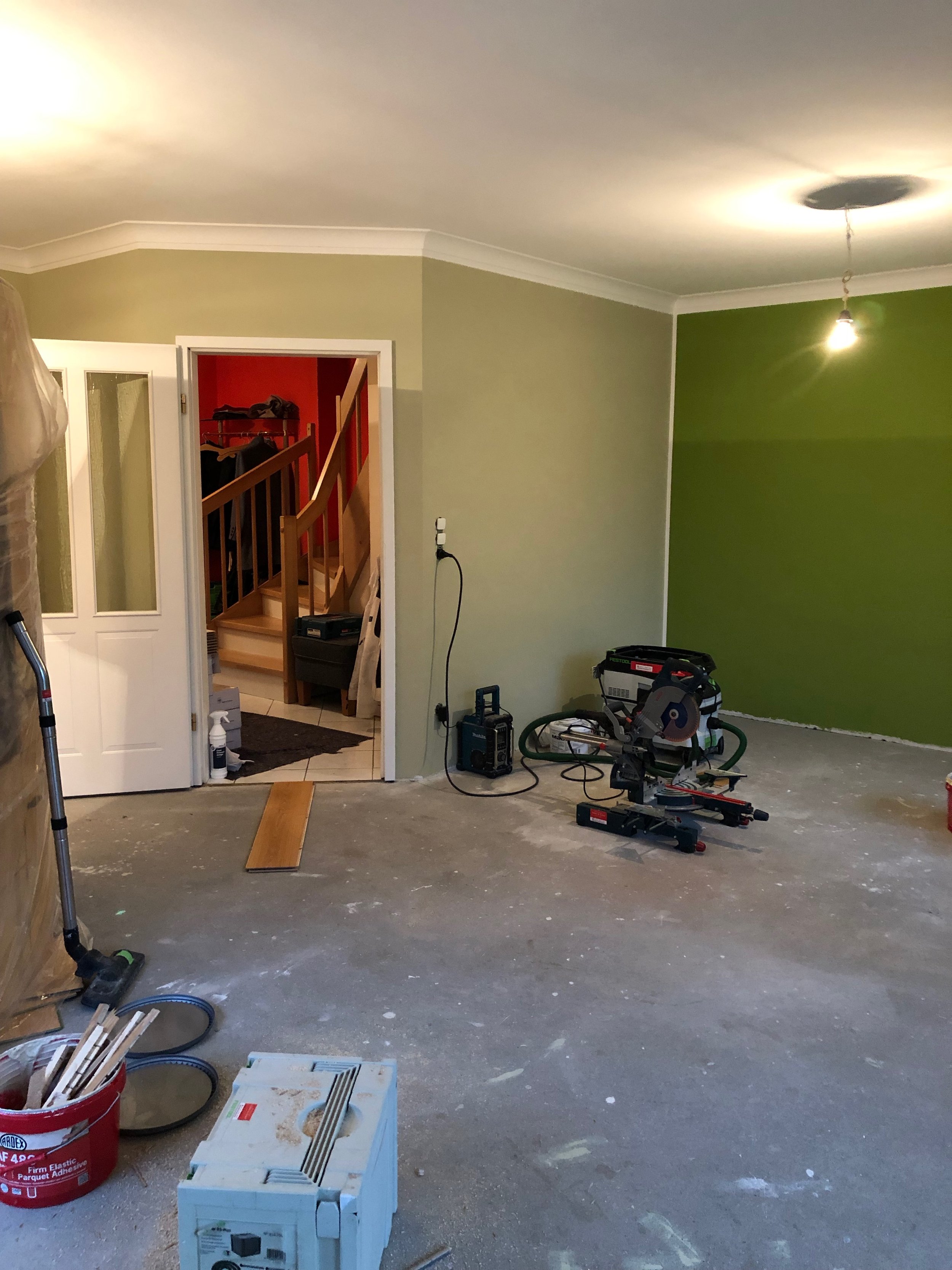 Ein Raum während Renovierungsarbeiten mit Werkzeugen wie einer Kreissäge, einem Staubsauger und Malerbedarf auf dem Boden, unvollständig gestrichene Wände in Grün und Beige, eine offene Tür zum Flur mit Treppe.