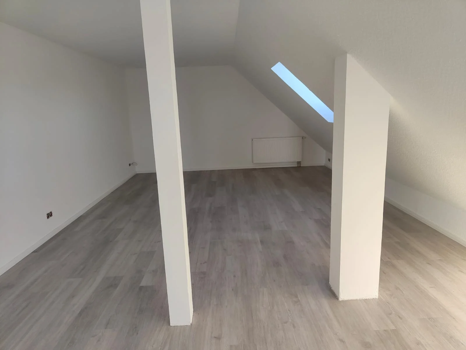 Ein leeres, helles Zimmer mit weißen Wänden, Holzboden und einer Dachschräge mit einem Dachfenster.