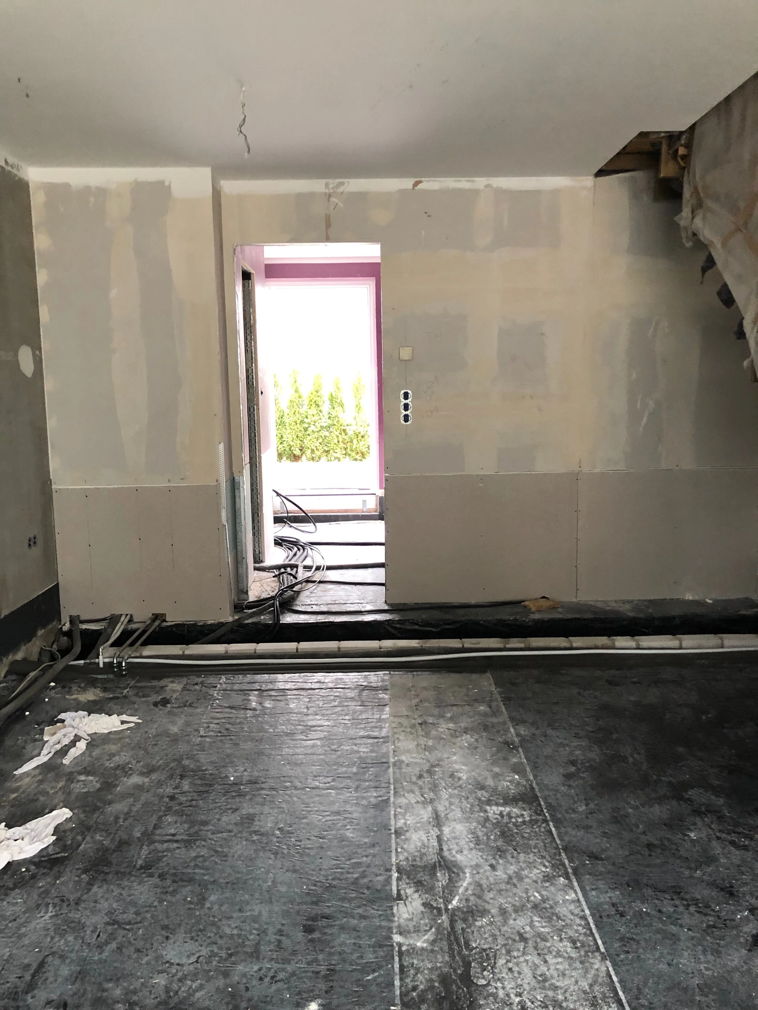 Innenaufnahme eines im Bau befindlichen Zimmers mit unvollständigen Wänden, sichtbaren Kabeln und Baumaterialien auf dem Boden.