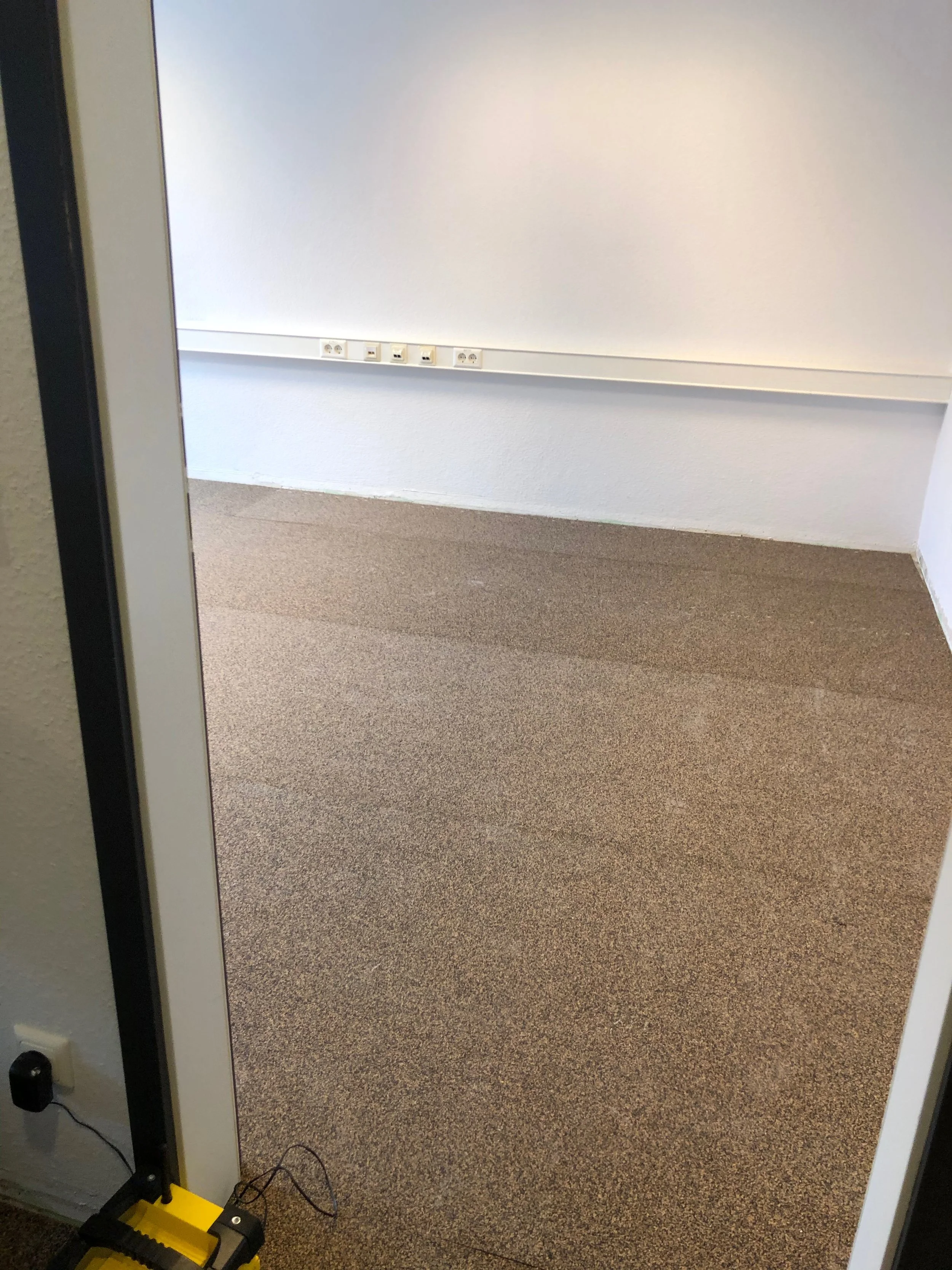 Leerer Büroraum mit weißen Wänden, braunem Teppichboden und mehreren Steckdosen an der Wand. Ein Kabel ist an einem kleinen, schwarzen und gelben Gerät angeschlossen, das auf dem Boden liegt.